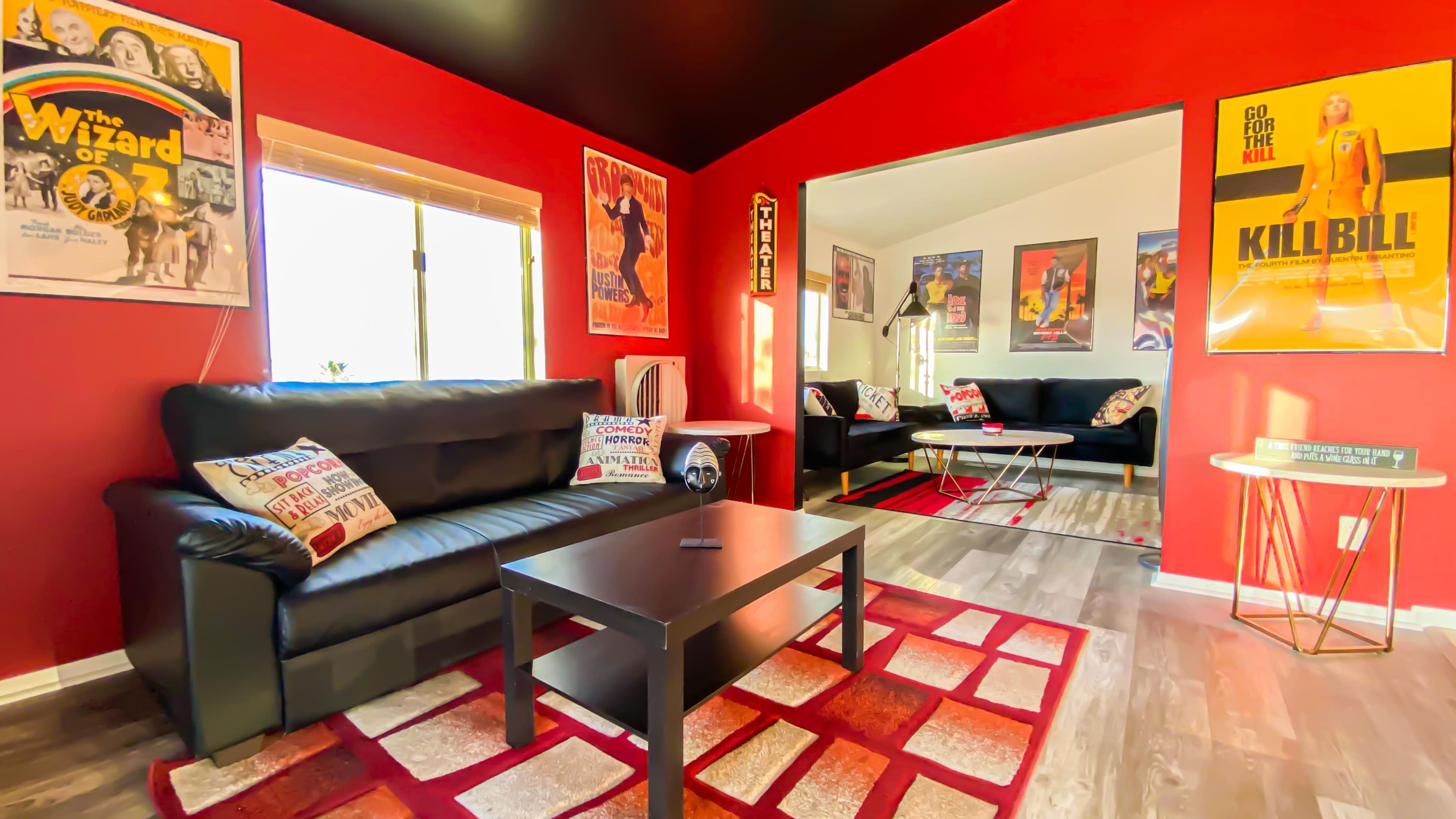 A living room with black leather sofas, a coffee table, and walls adorned with movie posters, set against a red color scheme.