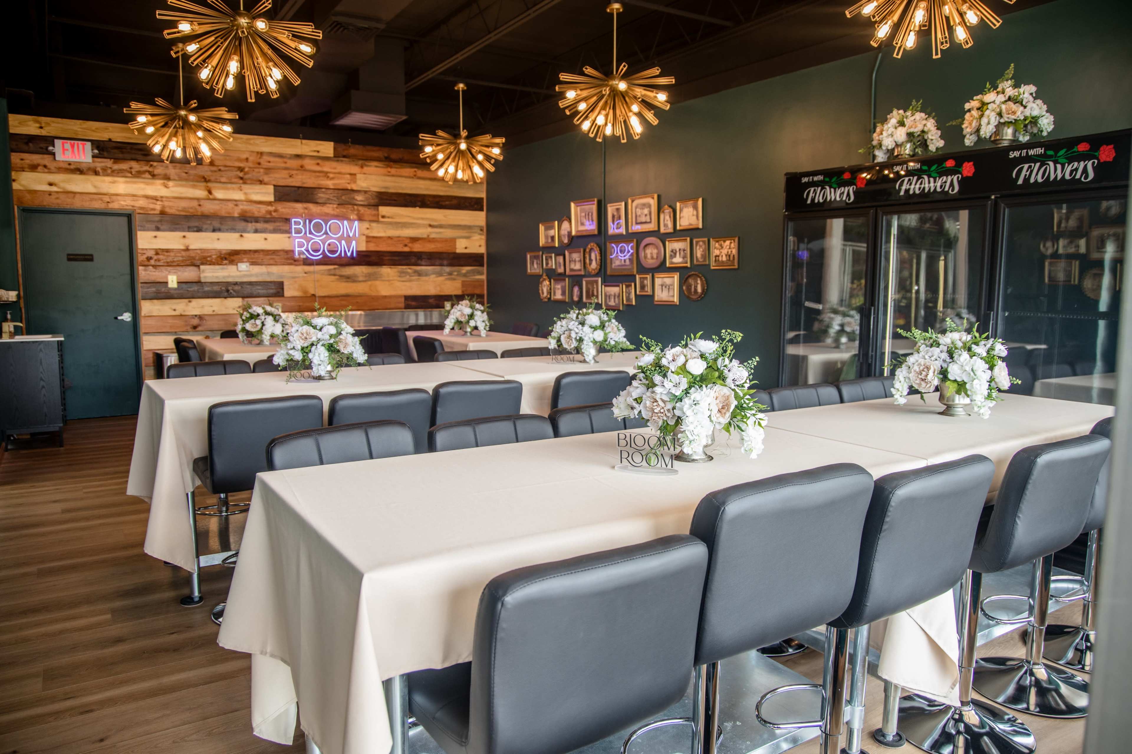 The image shows a stylish event space with multiple tables dressed in white tablecloths, decorated with flower arrangements, and a decorative wall featuring framed pictures and a neon sign that reads "BLOOM ROOM."