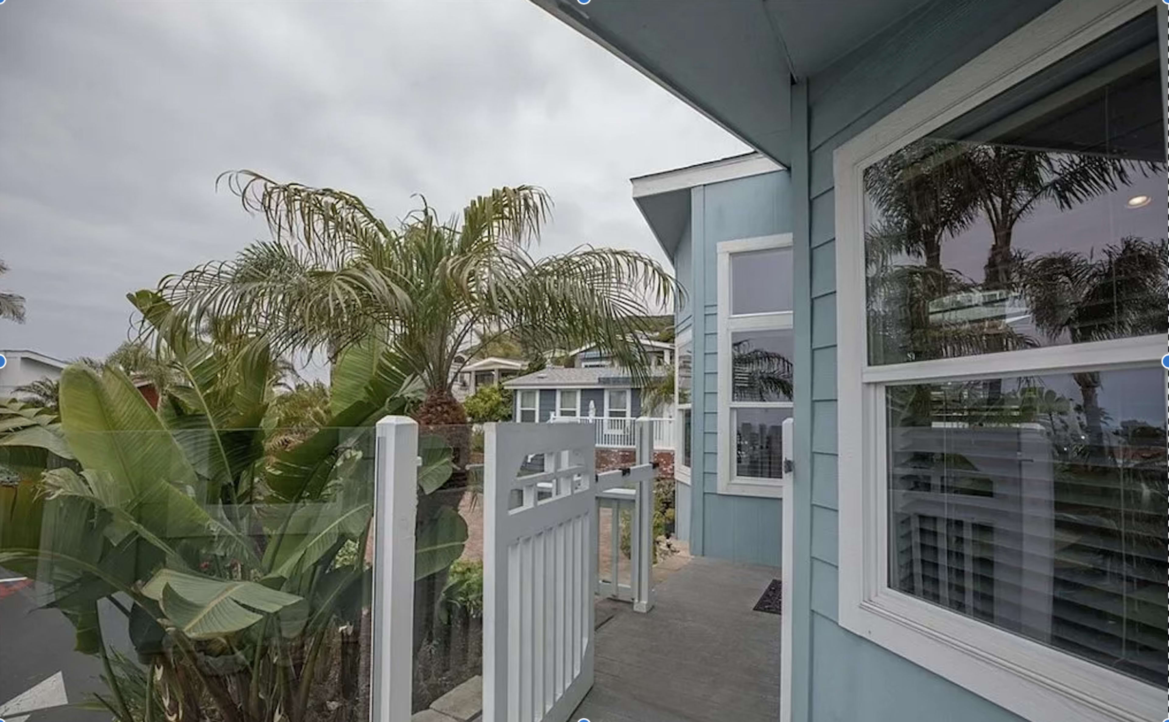 A blue house with palm trees and plants in the foreground, featuring large windows and a visible white fence.