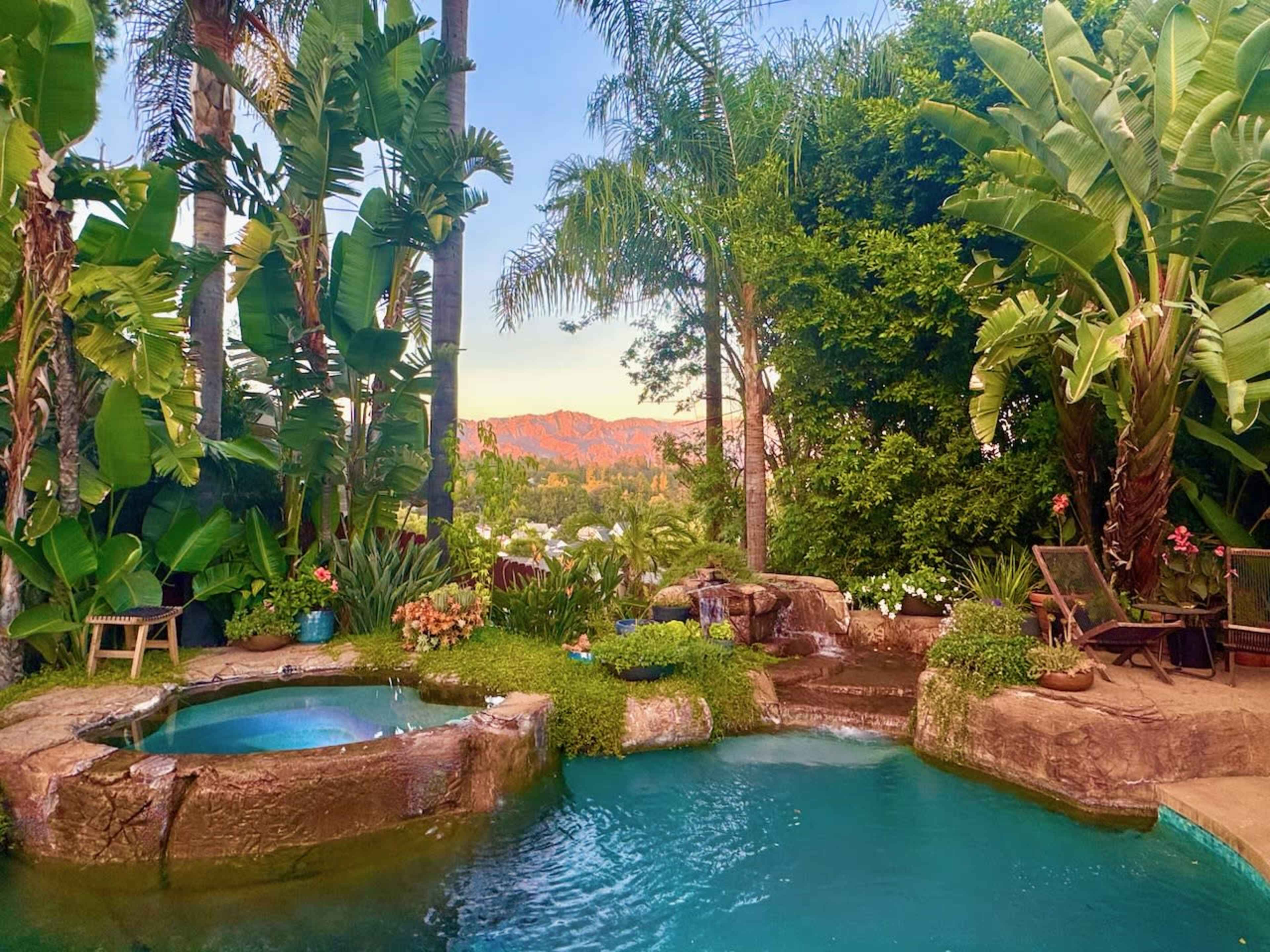 The image shows a landscaped backyard featuring a pool, a hot tub, lush green plants, and a view of distant mountains.