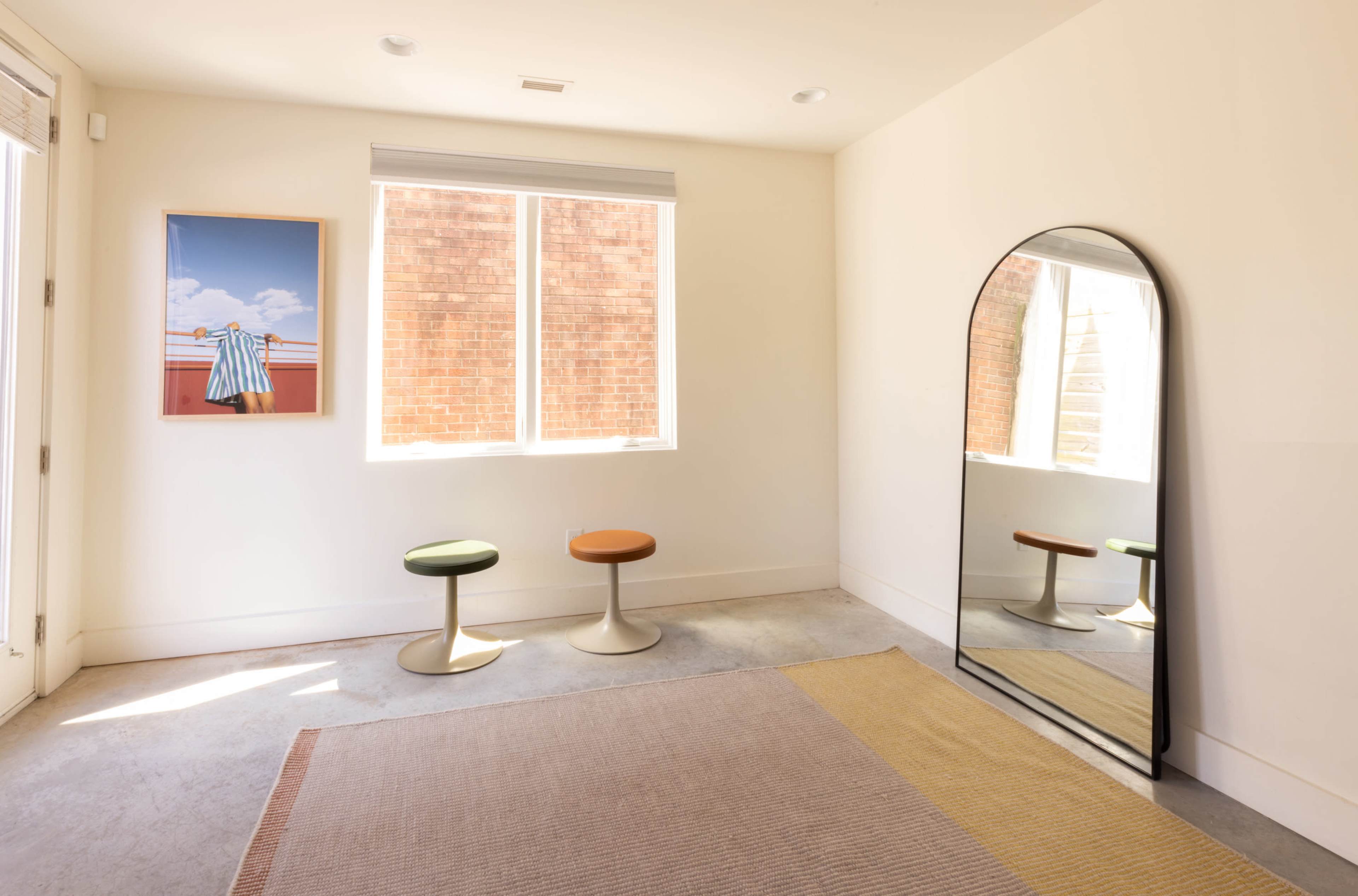 The room features a large mirror, two small stools in green and orange, a framed photograph on the wall, and natural light coming through a window.