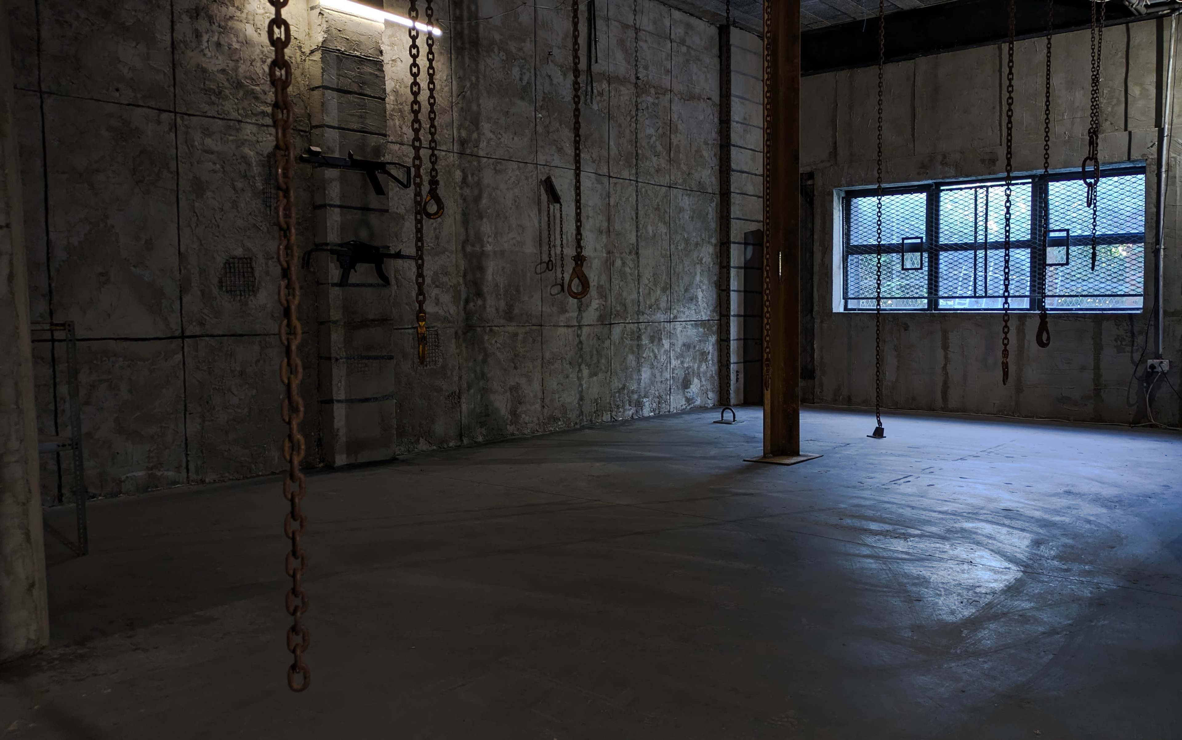 The image shows an industrial room with bare concrete walls, a metal pole in the center, and chains hanging from the ceiling.