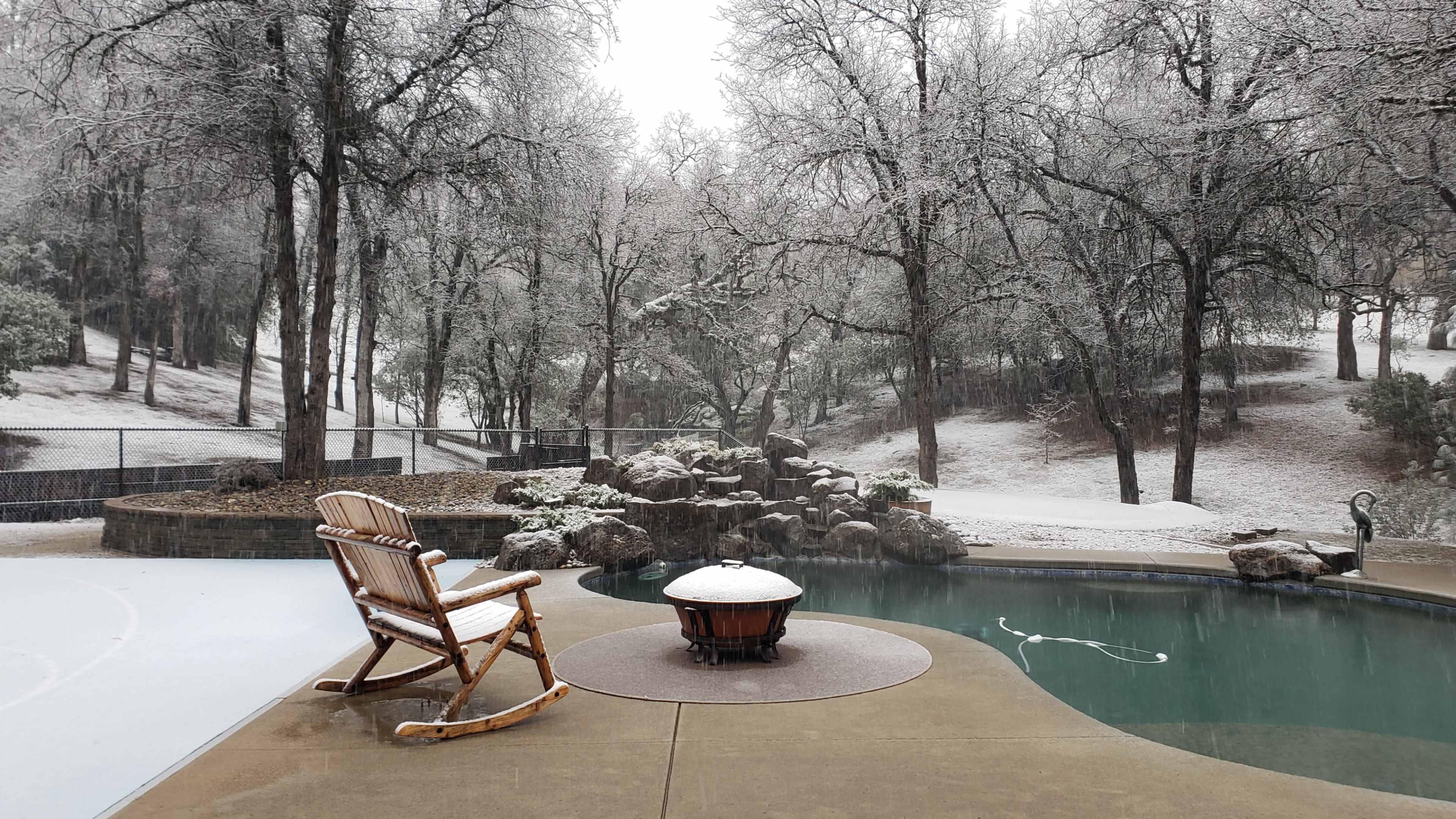 A wooden rocking chair sits near a snow-covered pool surrounded by trees.