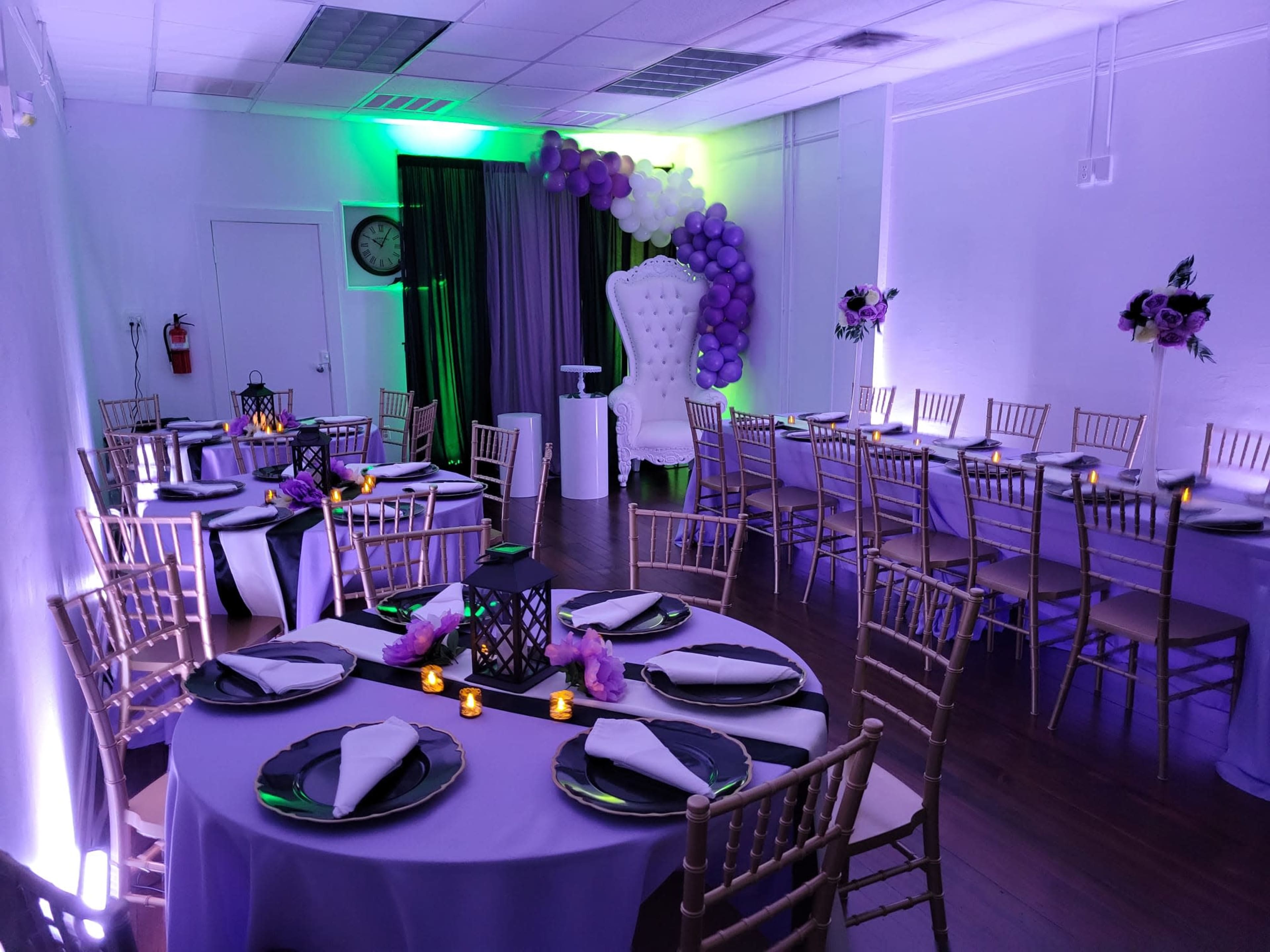 A banquet hall is elegantly decorated with purple and white balloons, tables set with black plates and napkins, and a large white chair at the front.