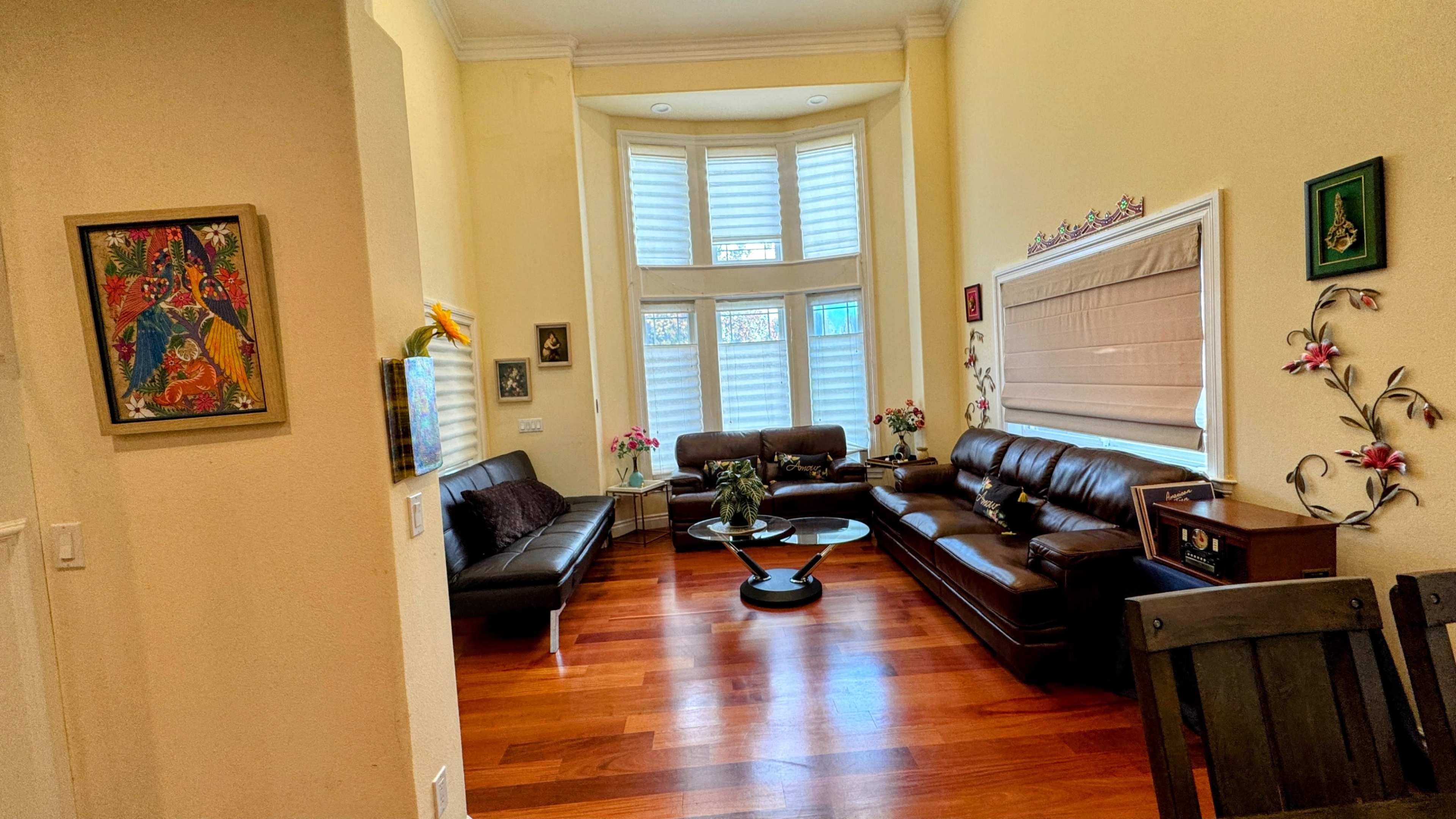 The image shows a spacious living room with hardwood floors, featuring two dark leather sofas, a round coffee table, and decorative elements on the walls.