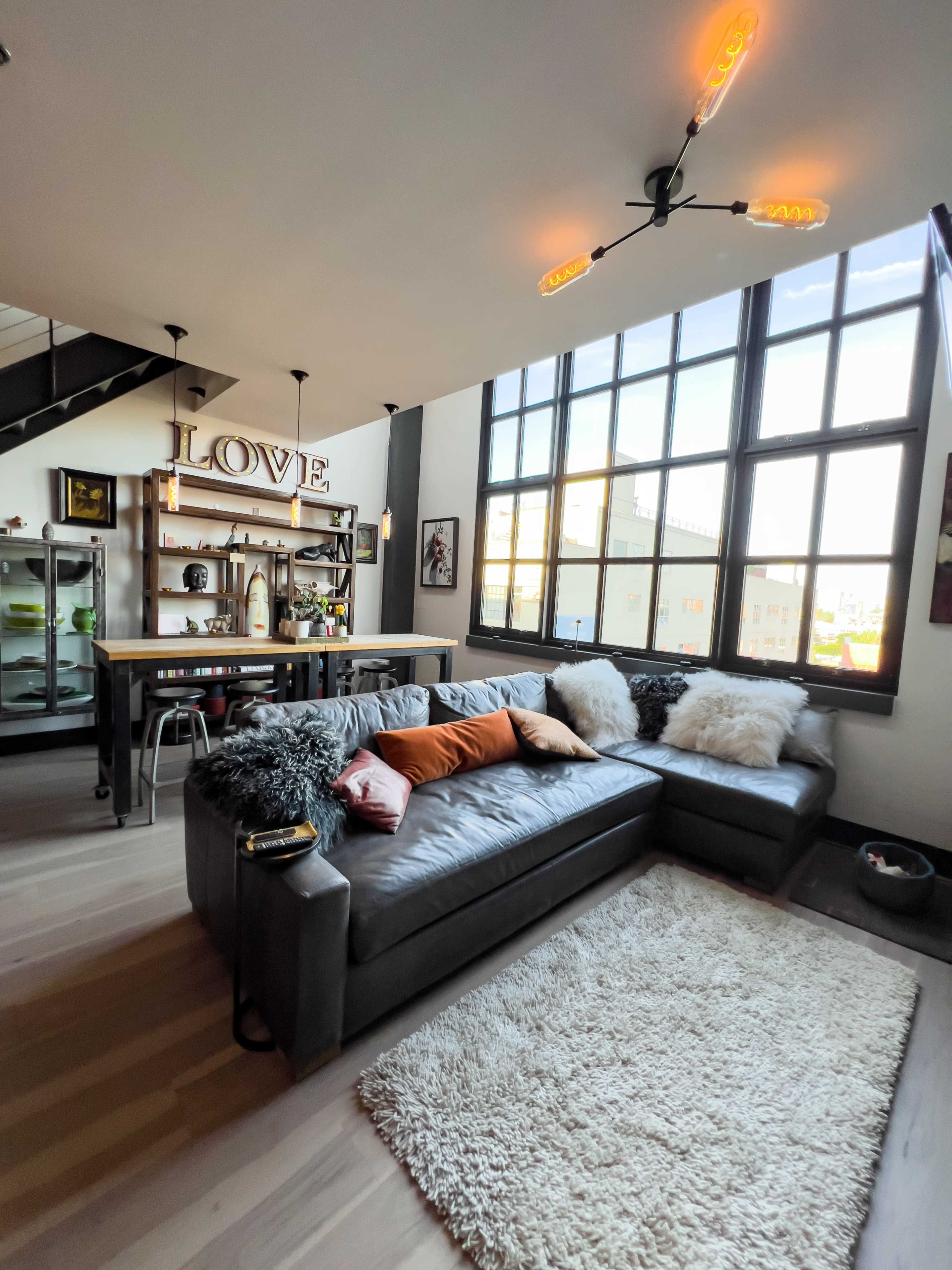 The image shows a modern living room featuring a large gray sectional sofa, a decorative rug, an industrial-style bar area, and large windows allowing natural light to enter the space.