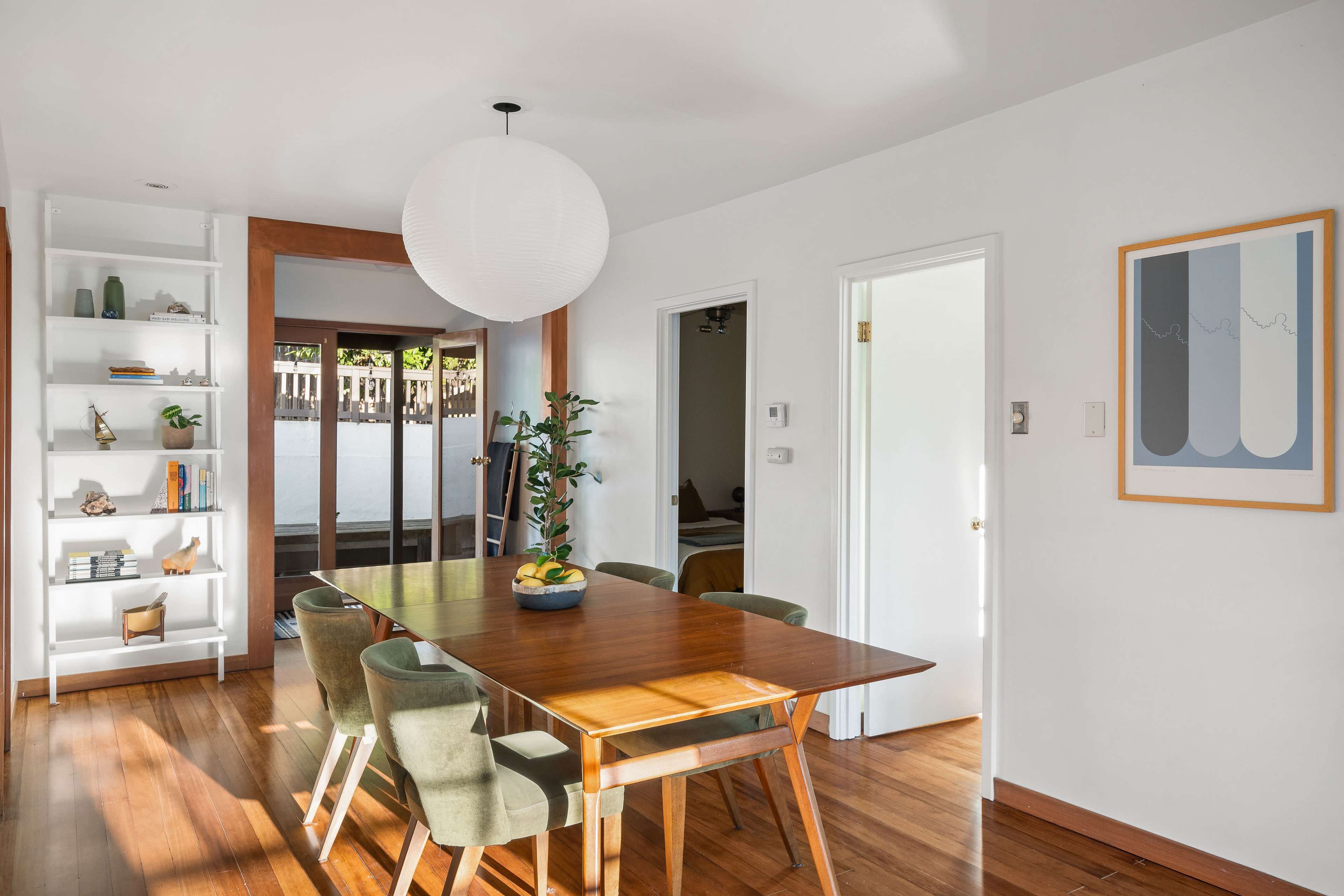 A dining area with a wooden table surrounded by chairs, a round pendant light overhead, and a bookshelf on one wall.