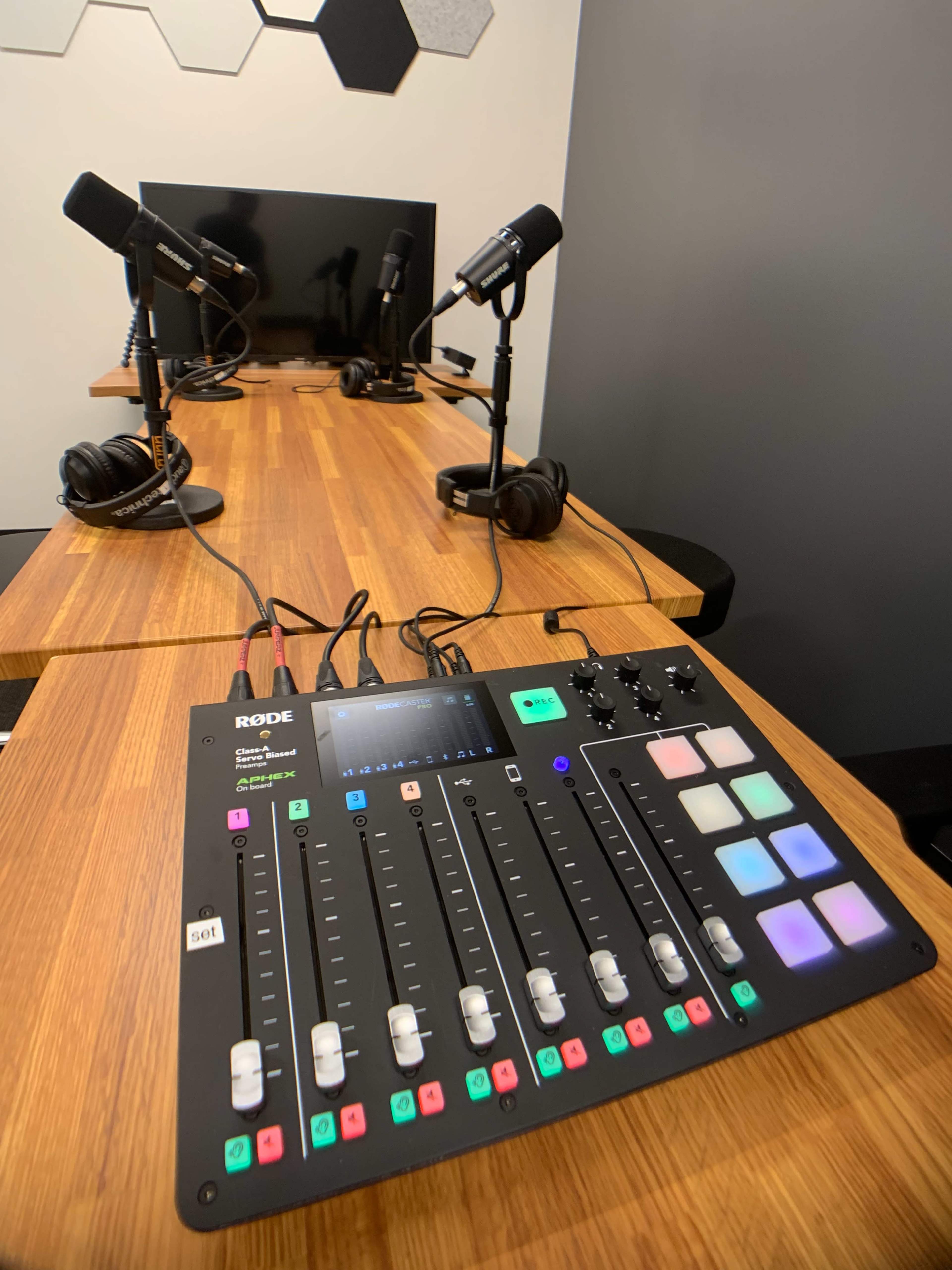 A wooden table with a Rode audio mixer, multiple microphones, and headphones set up in a recording studio.