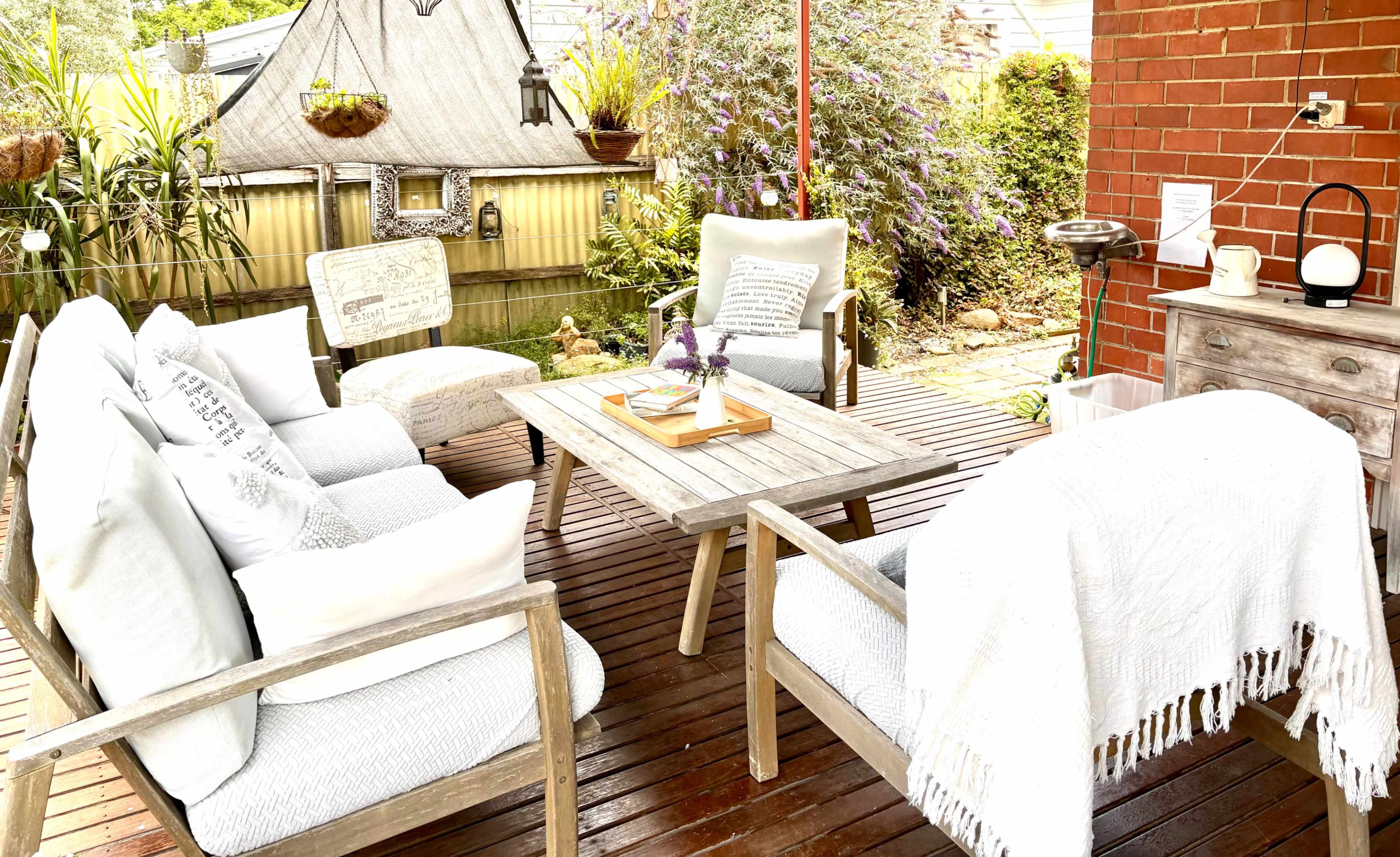 The image shows a cozy outdoor seating area with wooden furniture, including chairs and a table, surrounded by greenery and decorative plants.