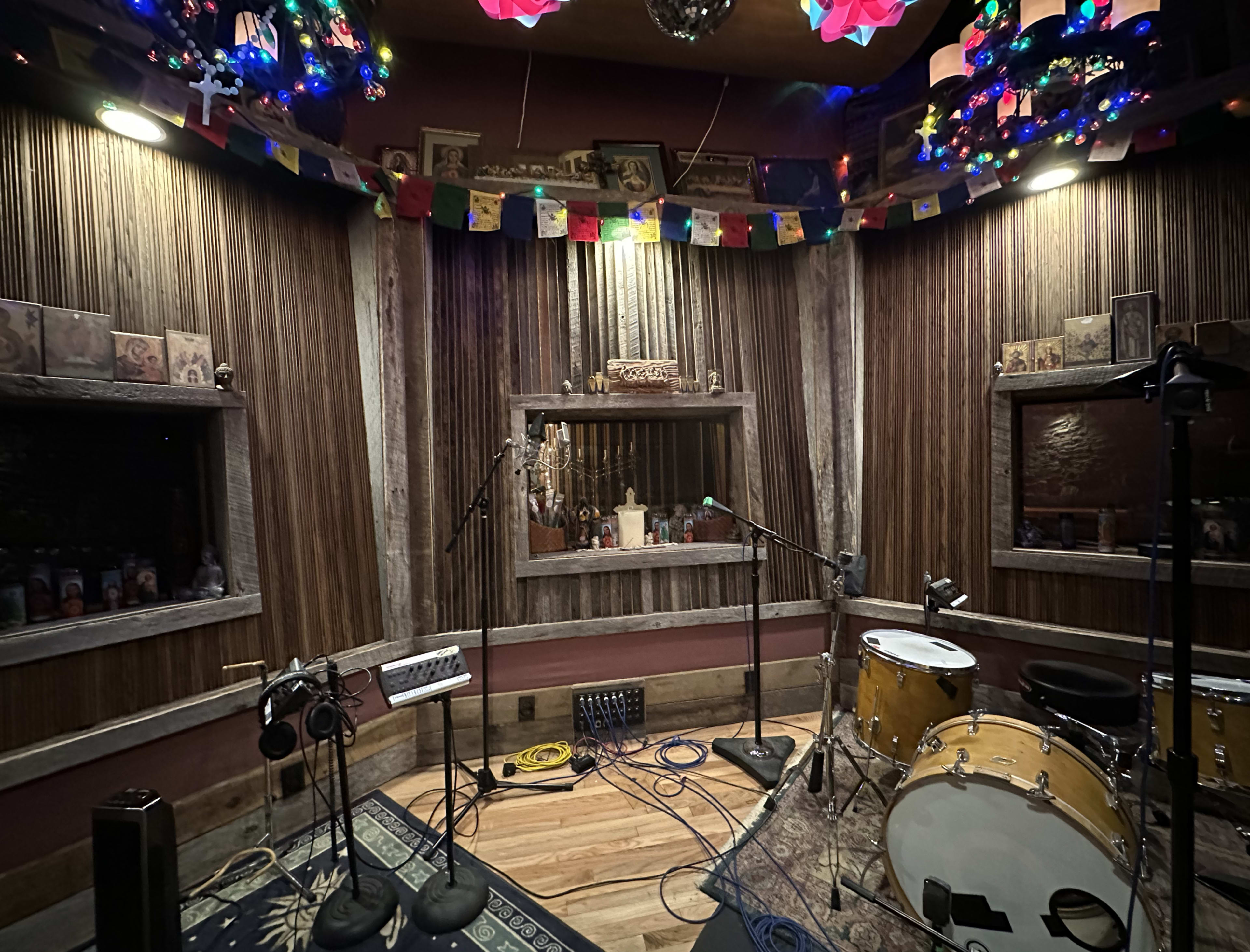The image shows a music recording studio with microphones, a drum set, and decorative elements like colorful lights and cultural artifacts on the walls.