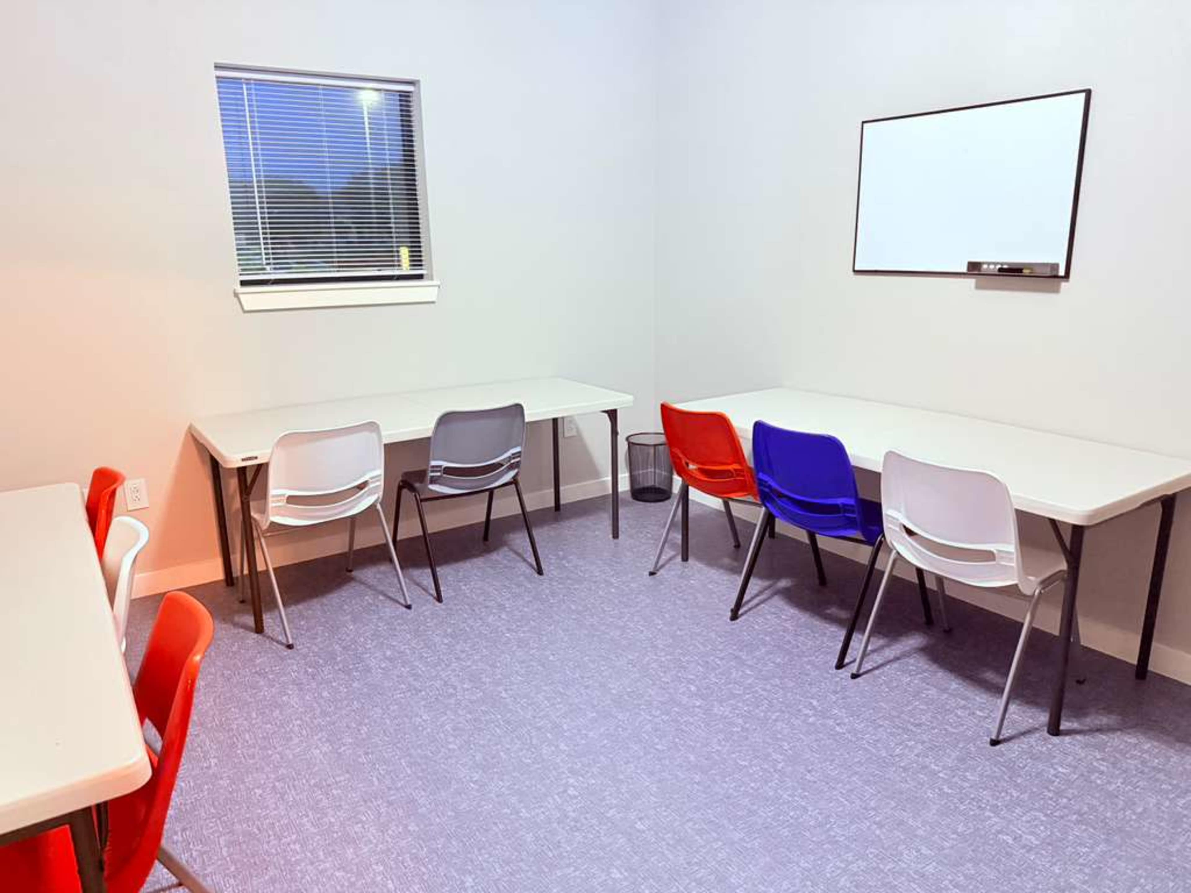 The image shows a small, well-lit classroom with two tables and a set of colorful chairs, along with a whiteboard mounted on the wall.