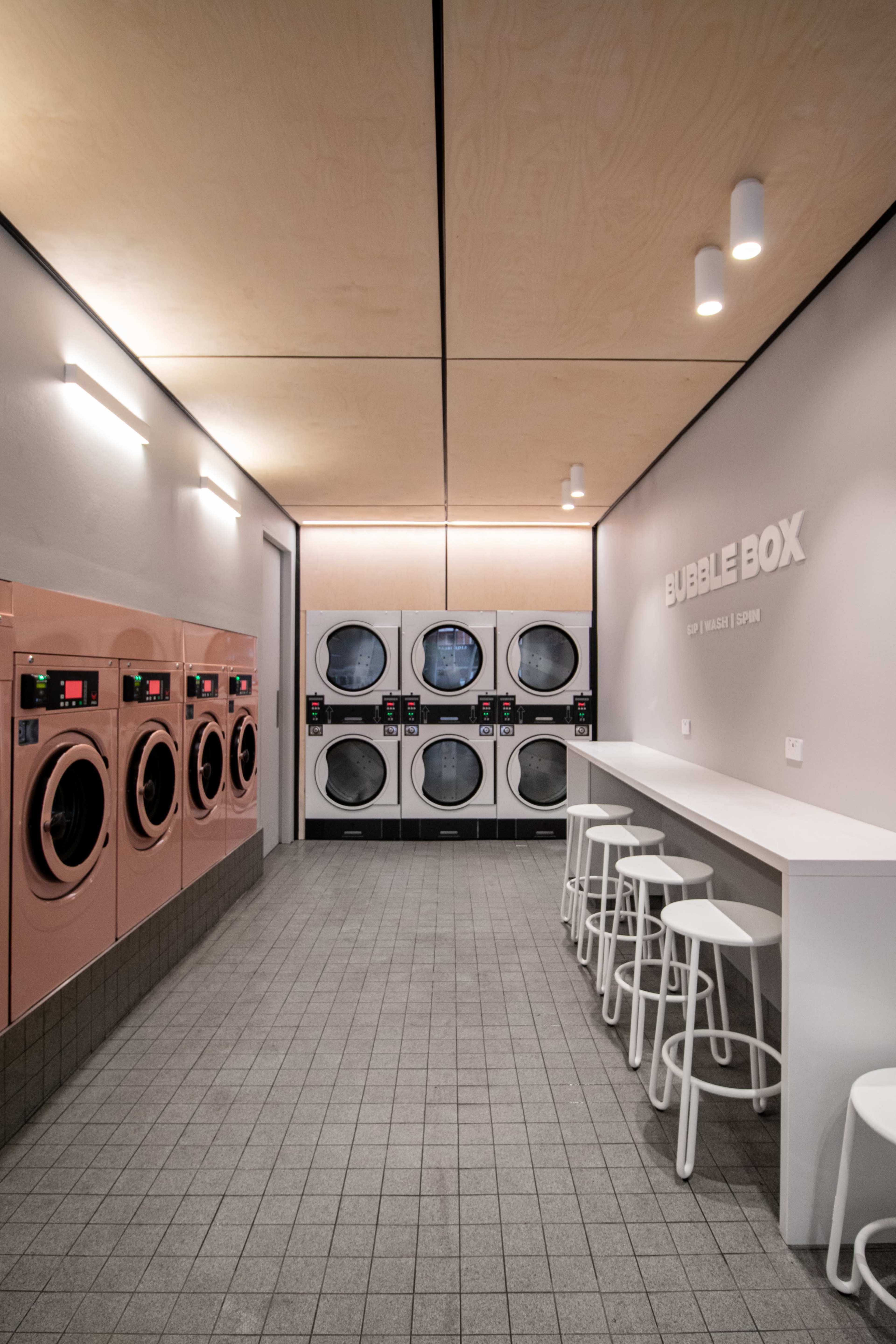 The interior of a modern laundromat features rows of washing machines and dryers against a minimalist wall, with stools and a counter for customers.