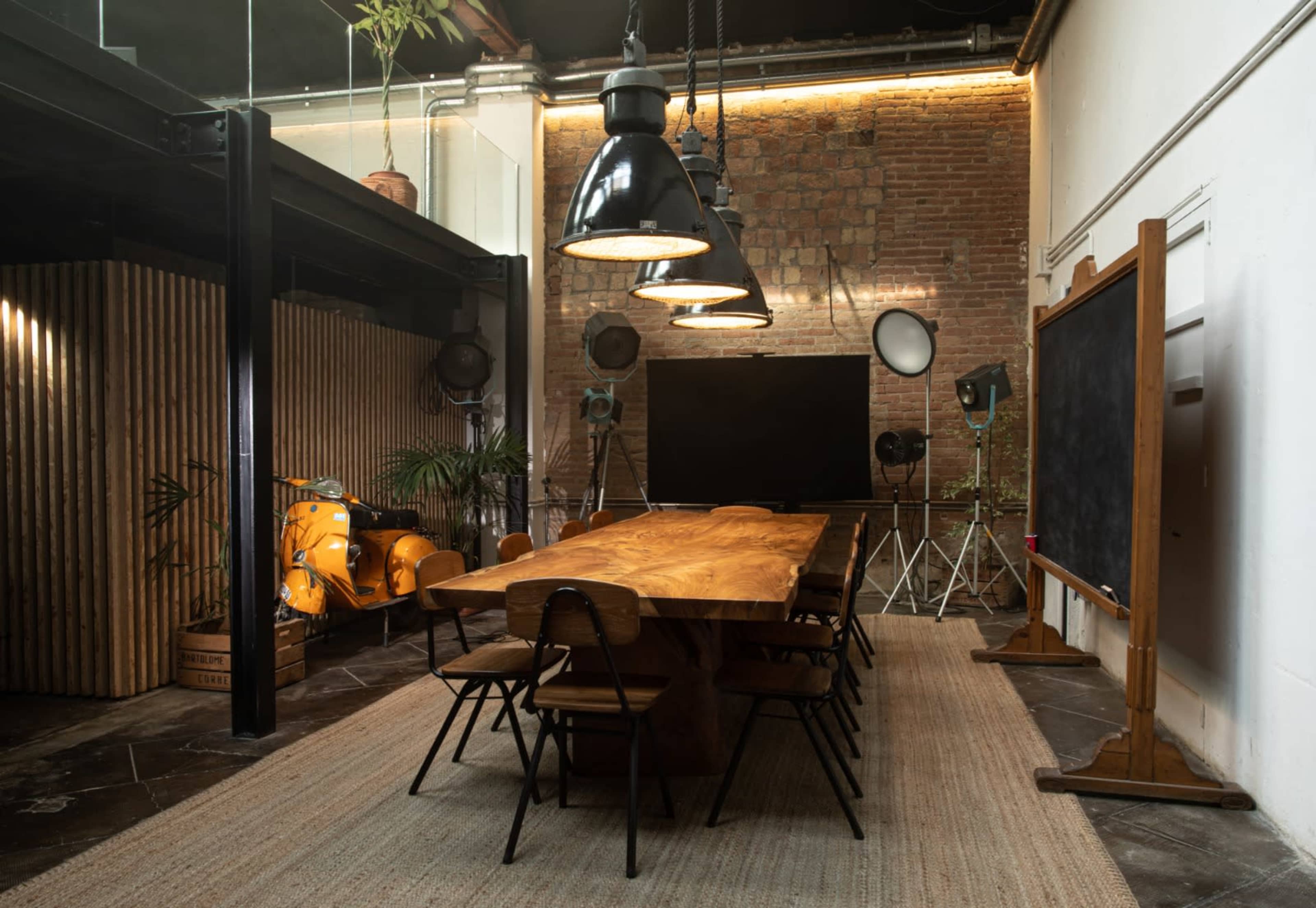 A long wooden dining table surrounded by black chairs is set in a spacious room featuring exposed brick walls and industrial lighting fixtures.