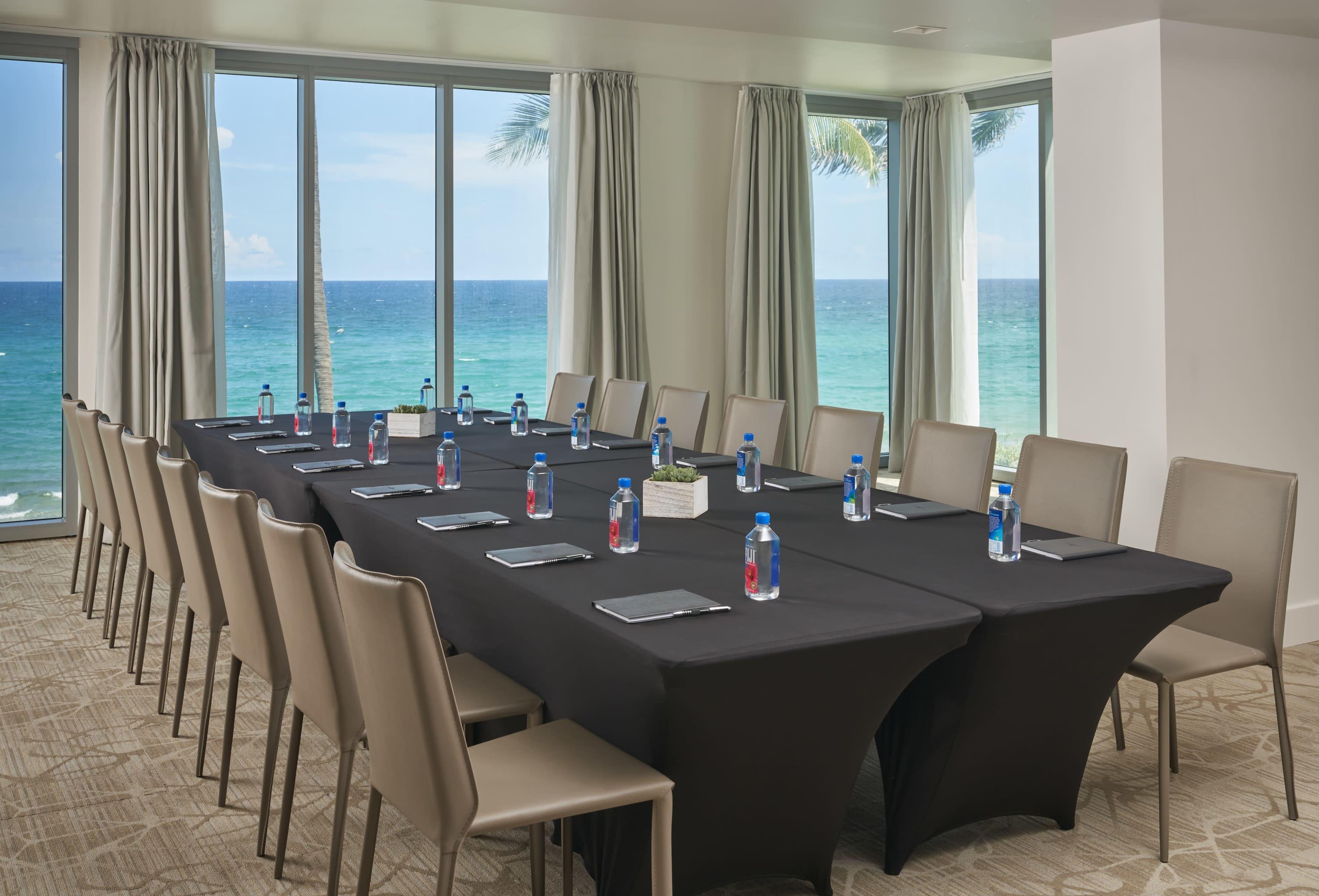 A long meeting table is set with water bottles and notepads, facing a large window that overlooks the ocean.