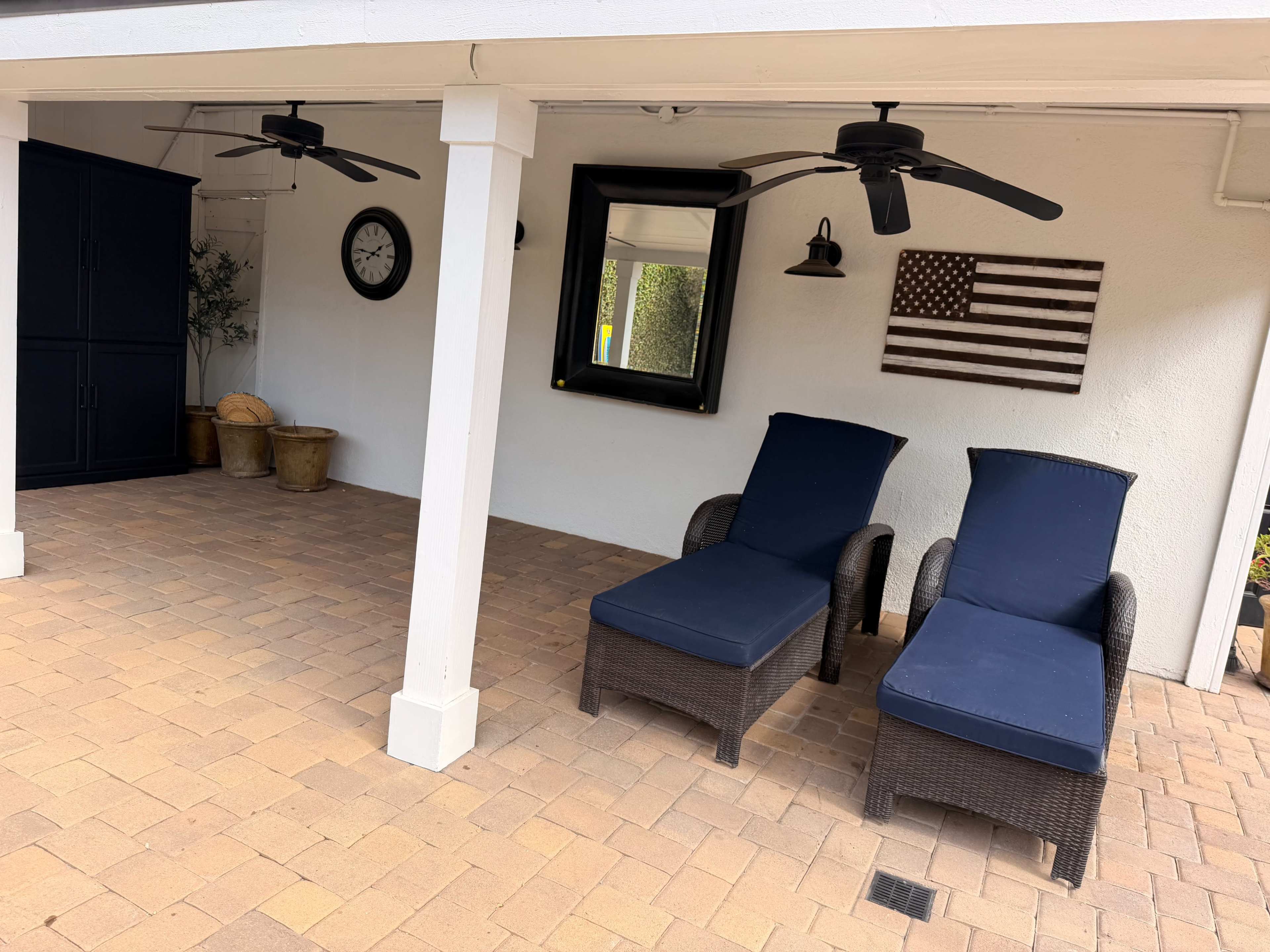 Two lounge chairs with blue cushions are positioned under a covered patio, next to a wall adorned with a clock and a decorative American flag.