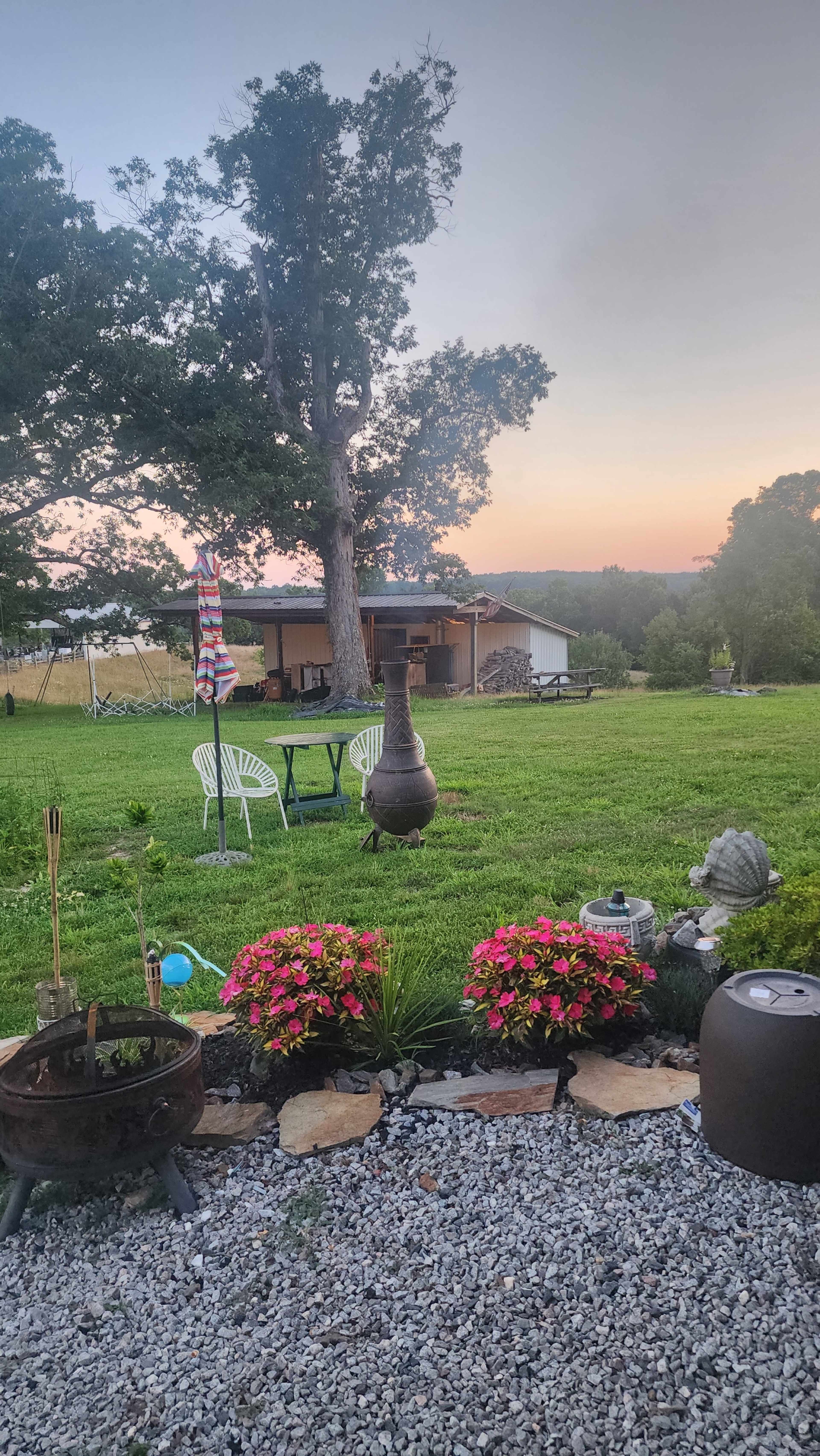 A green lawn with a table and chairs, a chiminea, and flower beds leads to a house with wood stacks and trees in the background at sunset.
