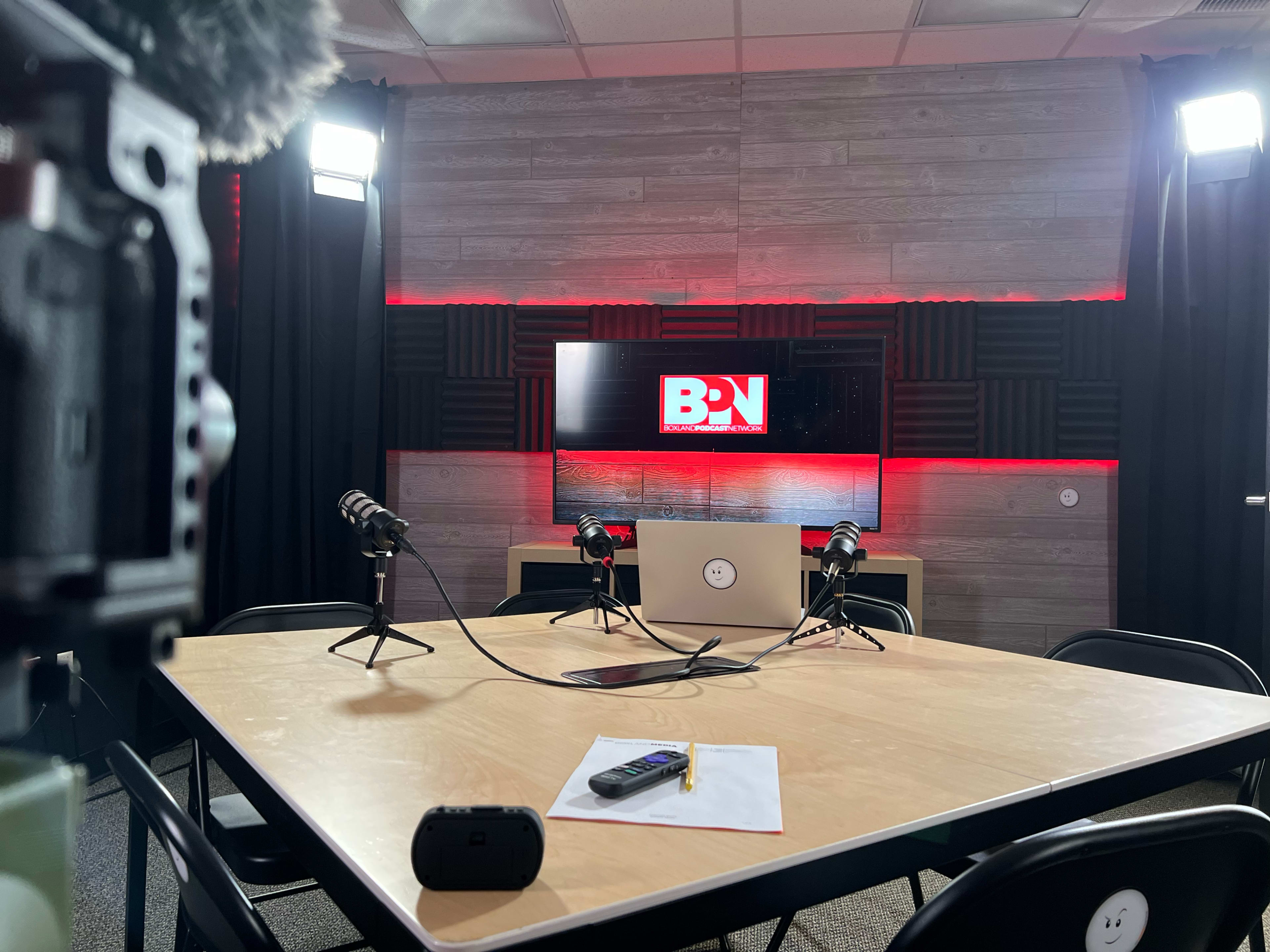 A modern studio setup features a round table with microphones, a laptop, and a monitor displaying a logo, surrounded by black curtains and bright lighting.