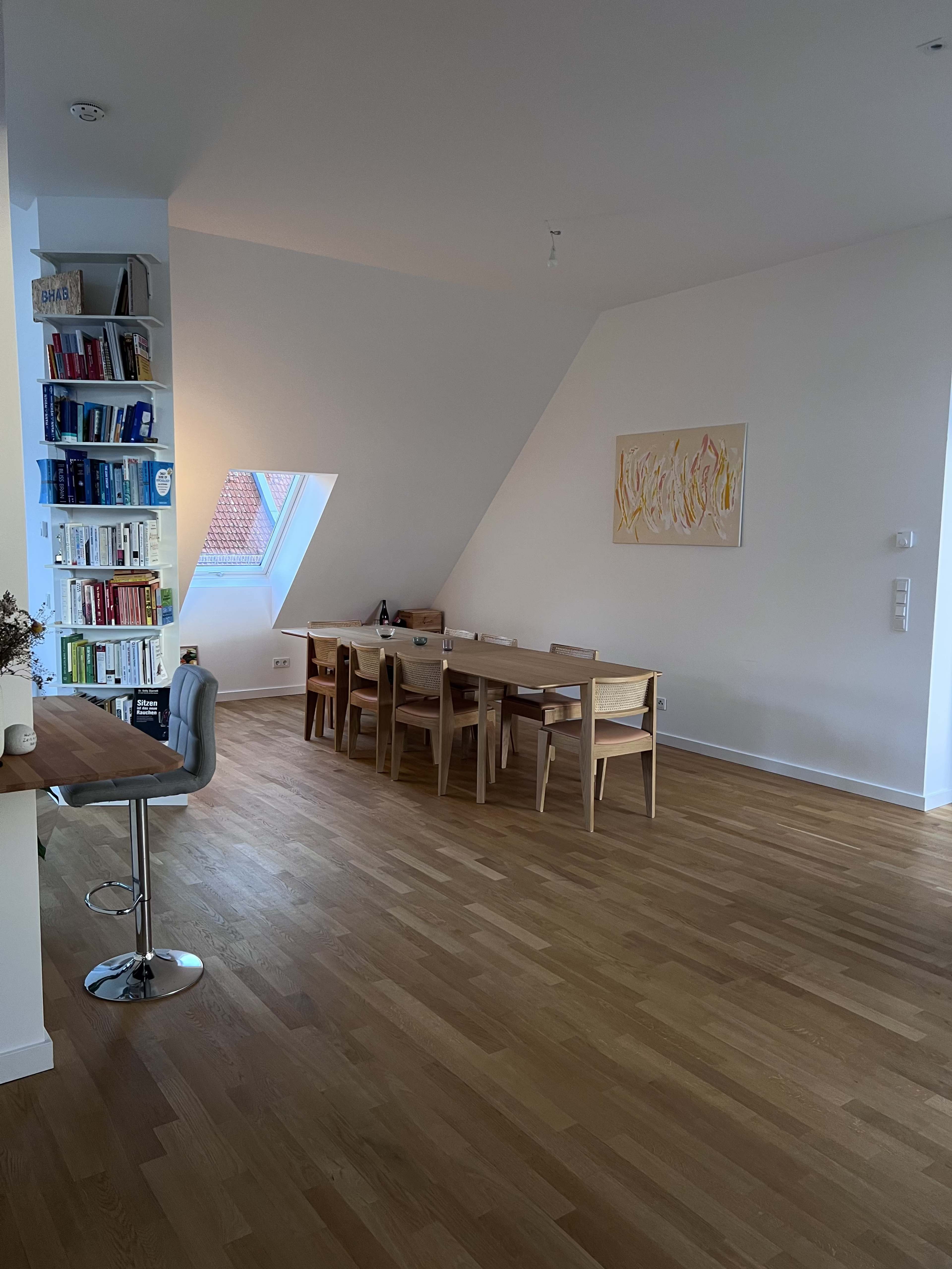 The image shows a modern dining area with a large wooden table surrounded by chairs, a tall bookshelf filled with books, and a slanted wall featuring a window.