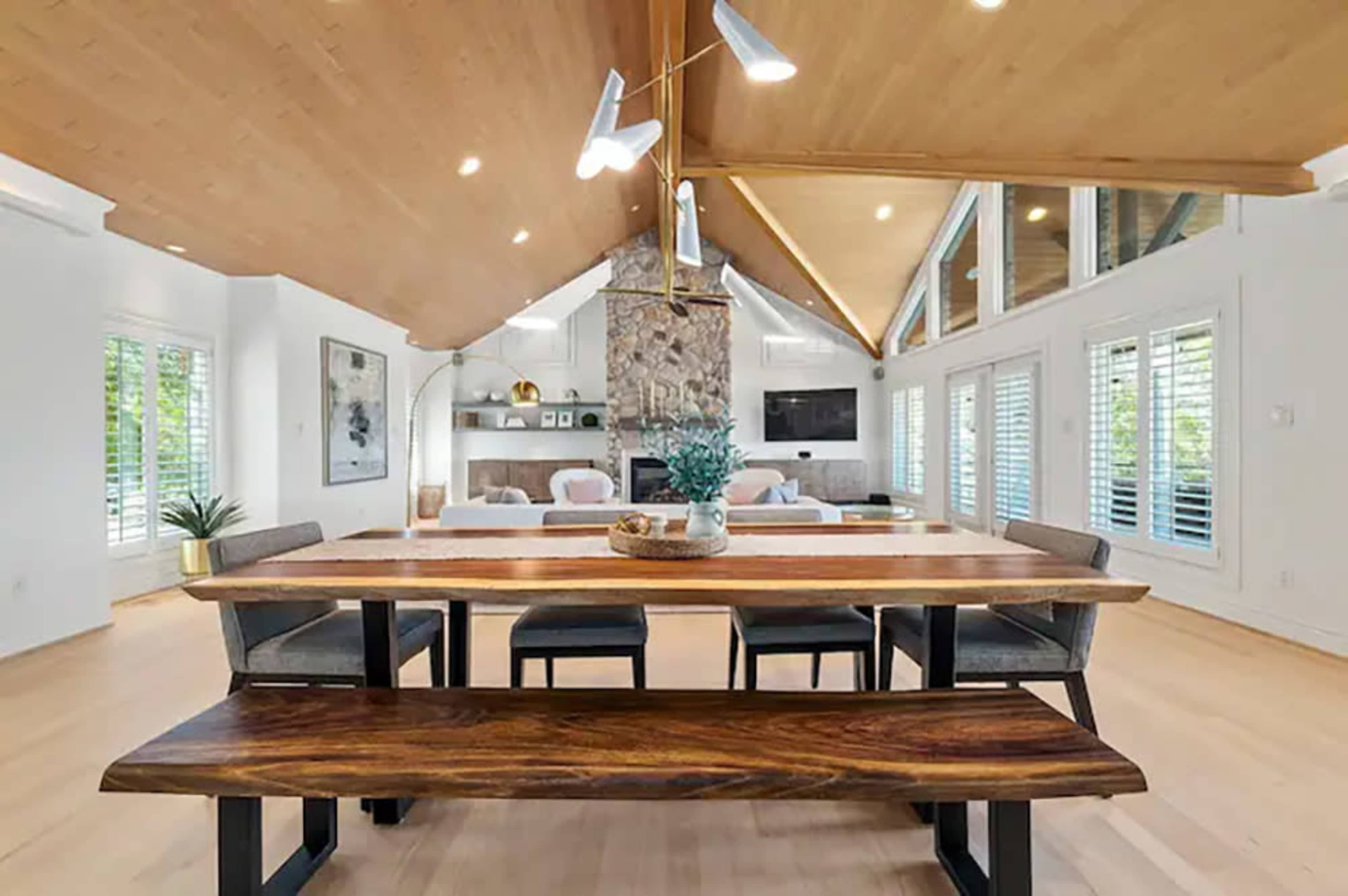 The image shows a modern dining area with a large wooden table and bench, surrounded by chairs, and featuring a stone fireplace and large windows with greenery visible outside.