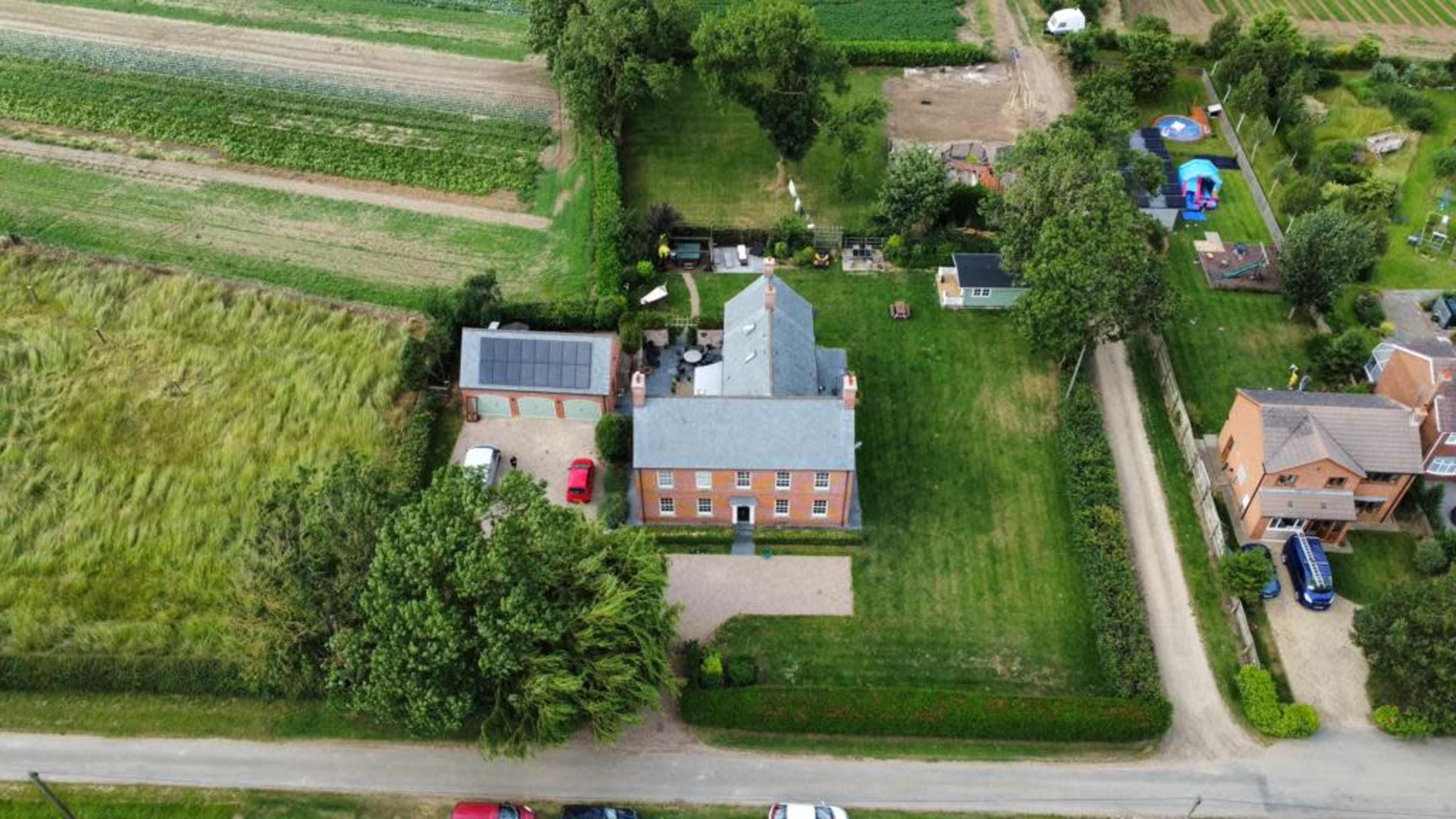 An aerial view of a rural property featuring a large house surrounded by lawns and fields, with several vehicles parked in the driveway.