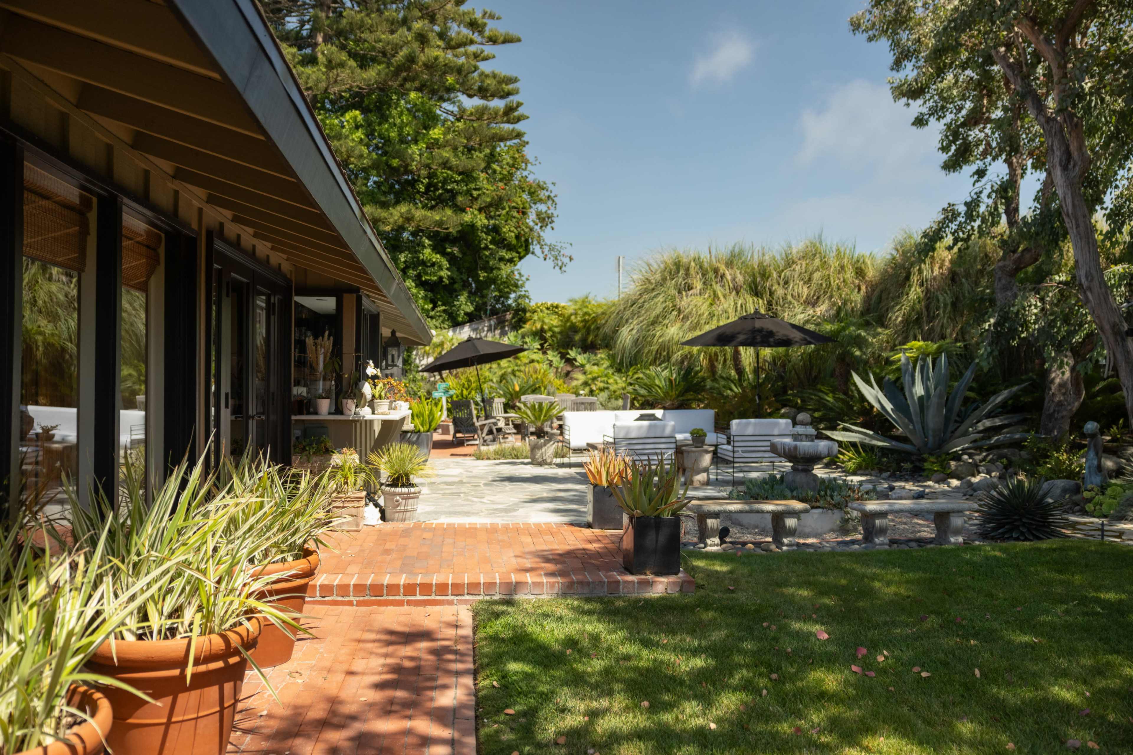 A modern patio area features landscaped gardens with various plants, dining tables under umbrellas, and a stone pathway leading to a seating area.