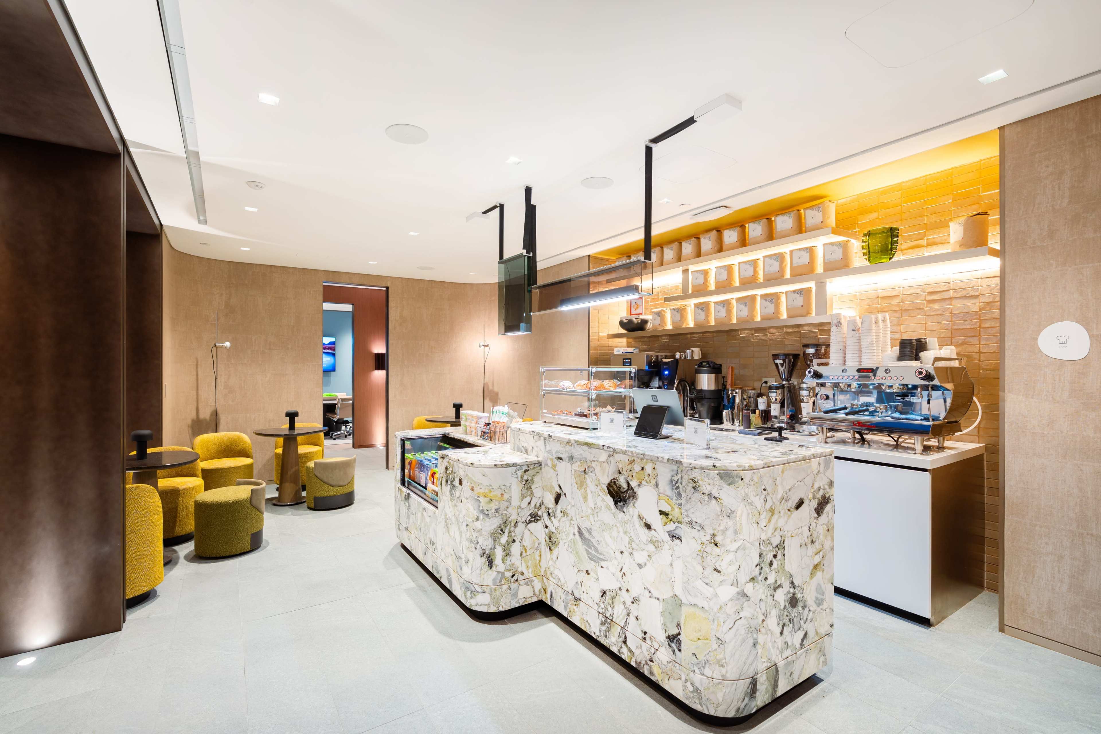 The image shows a modern café with a marble counter, a coffee machine, and seating areas featuring yellow and green chairs.