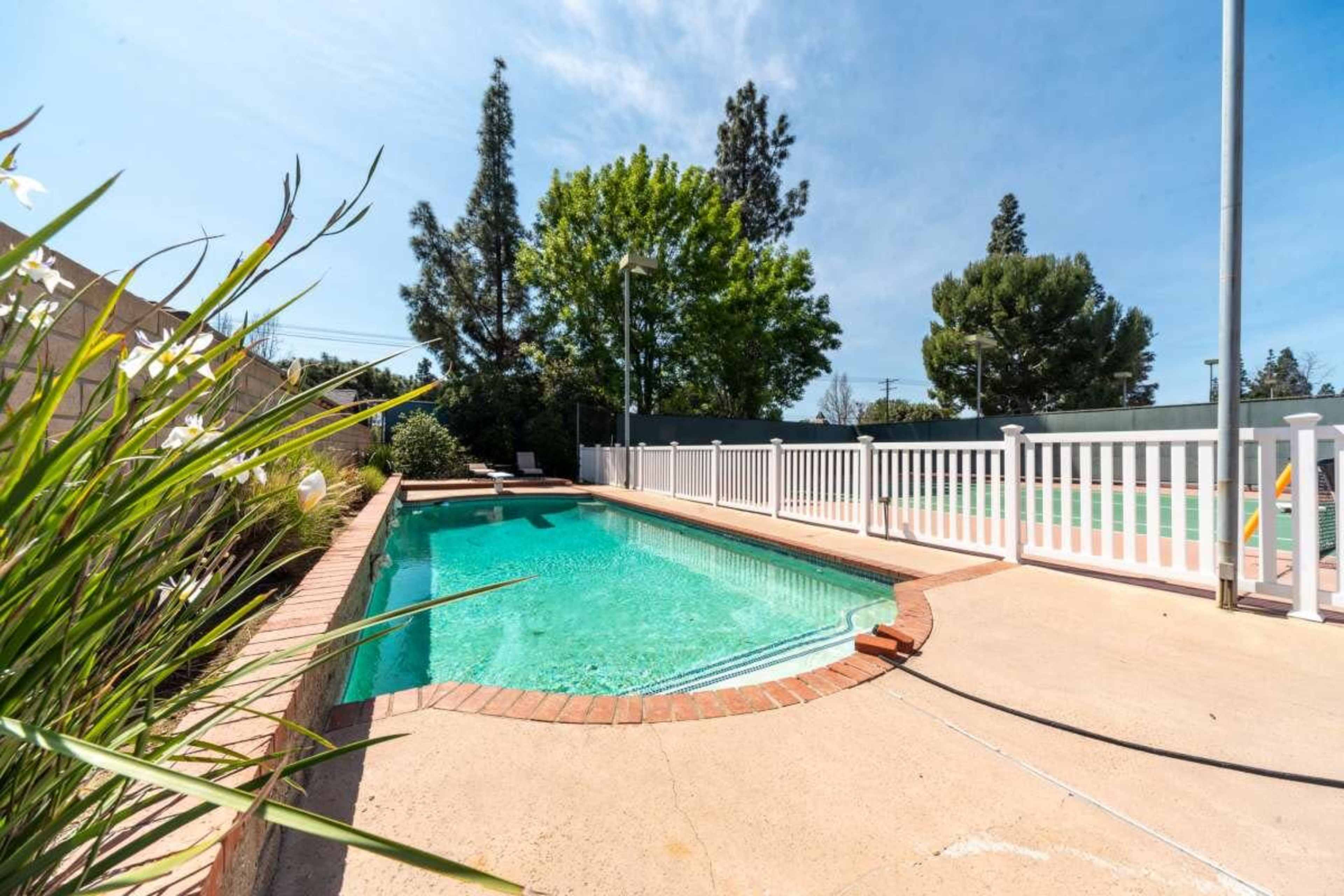 The image shows a clear swimming pool surrounded by a concrete deck and a white railing, with trees and greenery in the background.