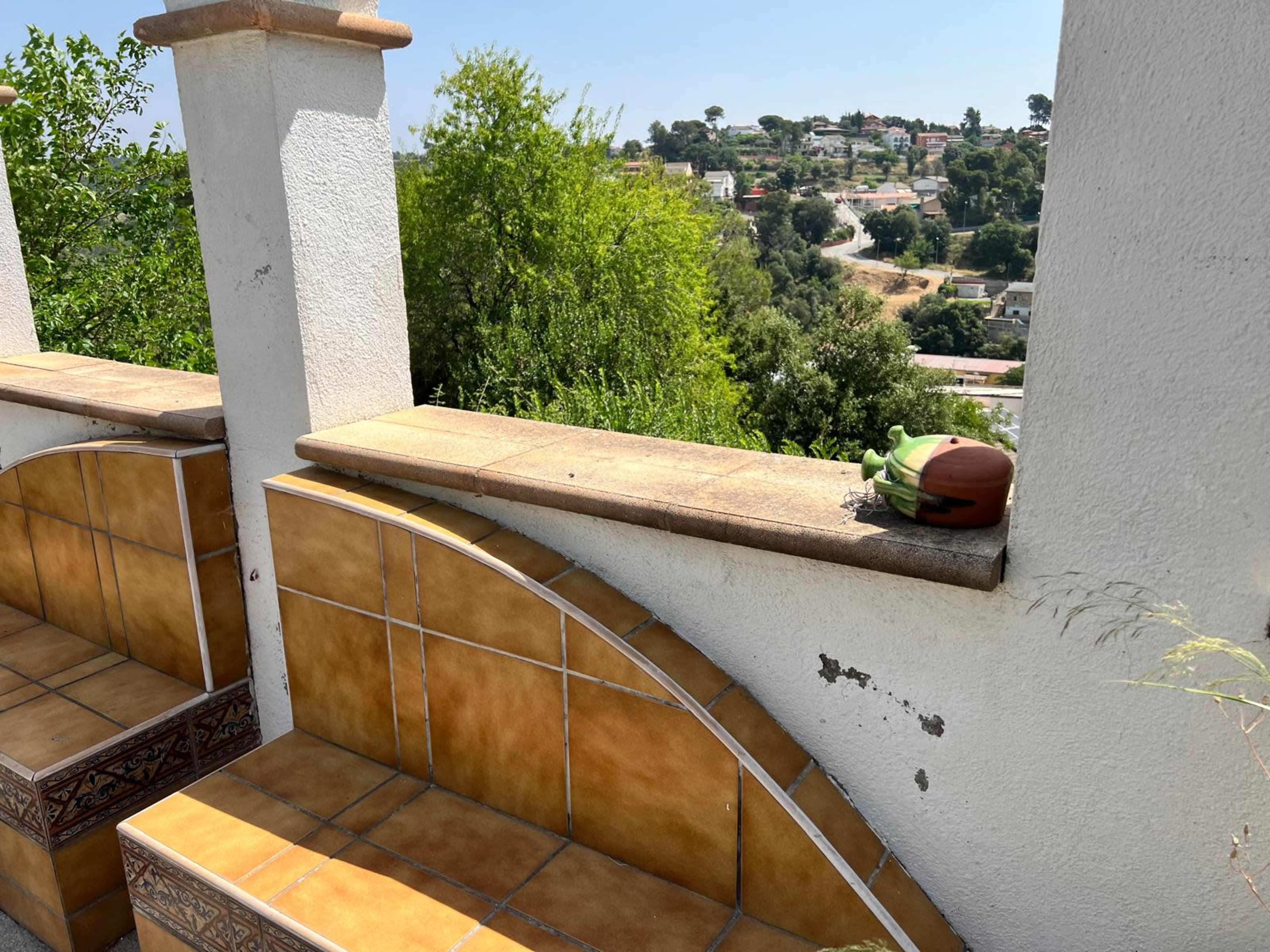 A green object and a small brown item rest on a tiled ledge with a view of a tree-filled hillside and distant houses.