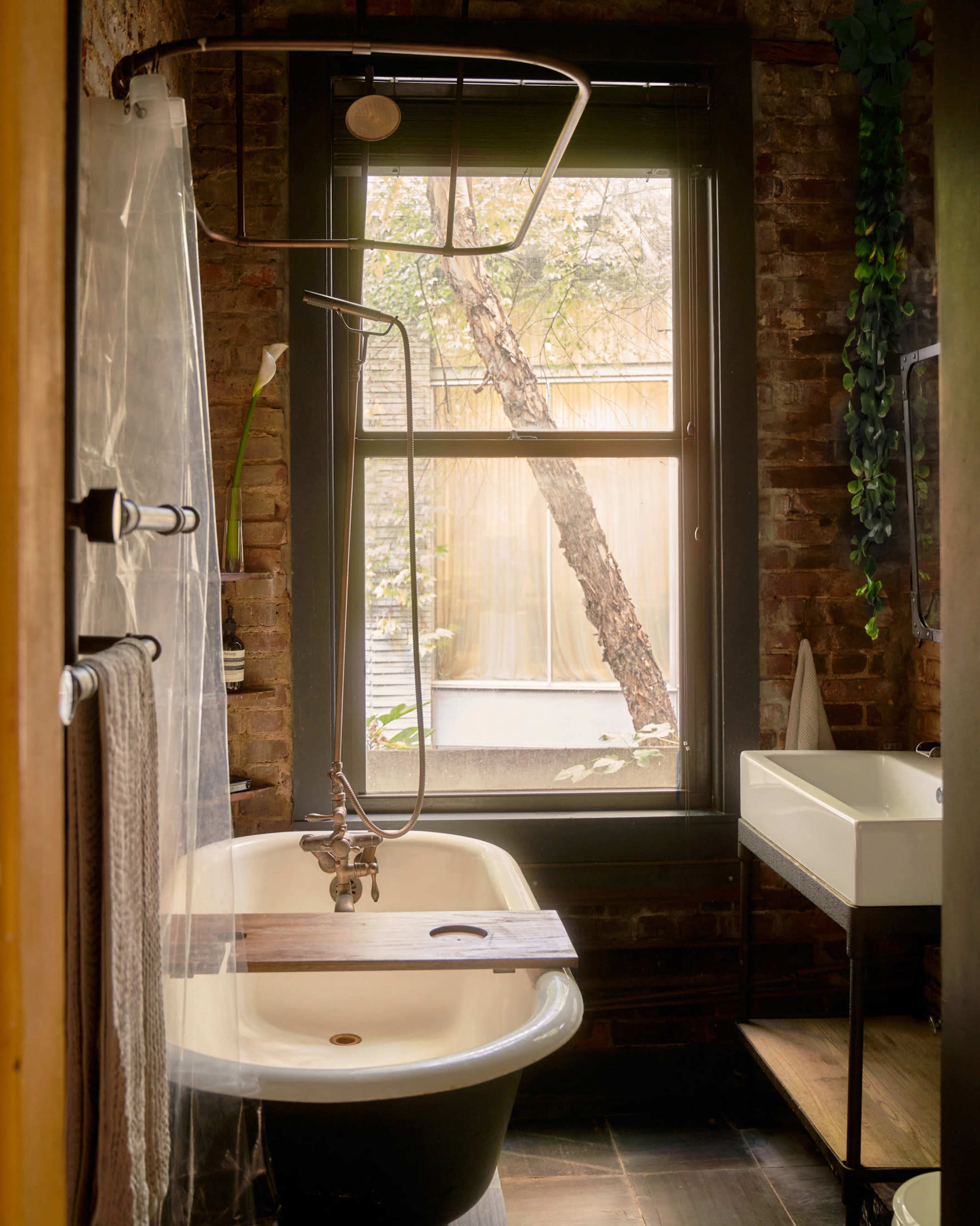 A vintage bathtub is positioned near a large window, framed by exposed brick walls and decorated with a wooden shower rod and a small sink.
