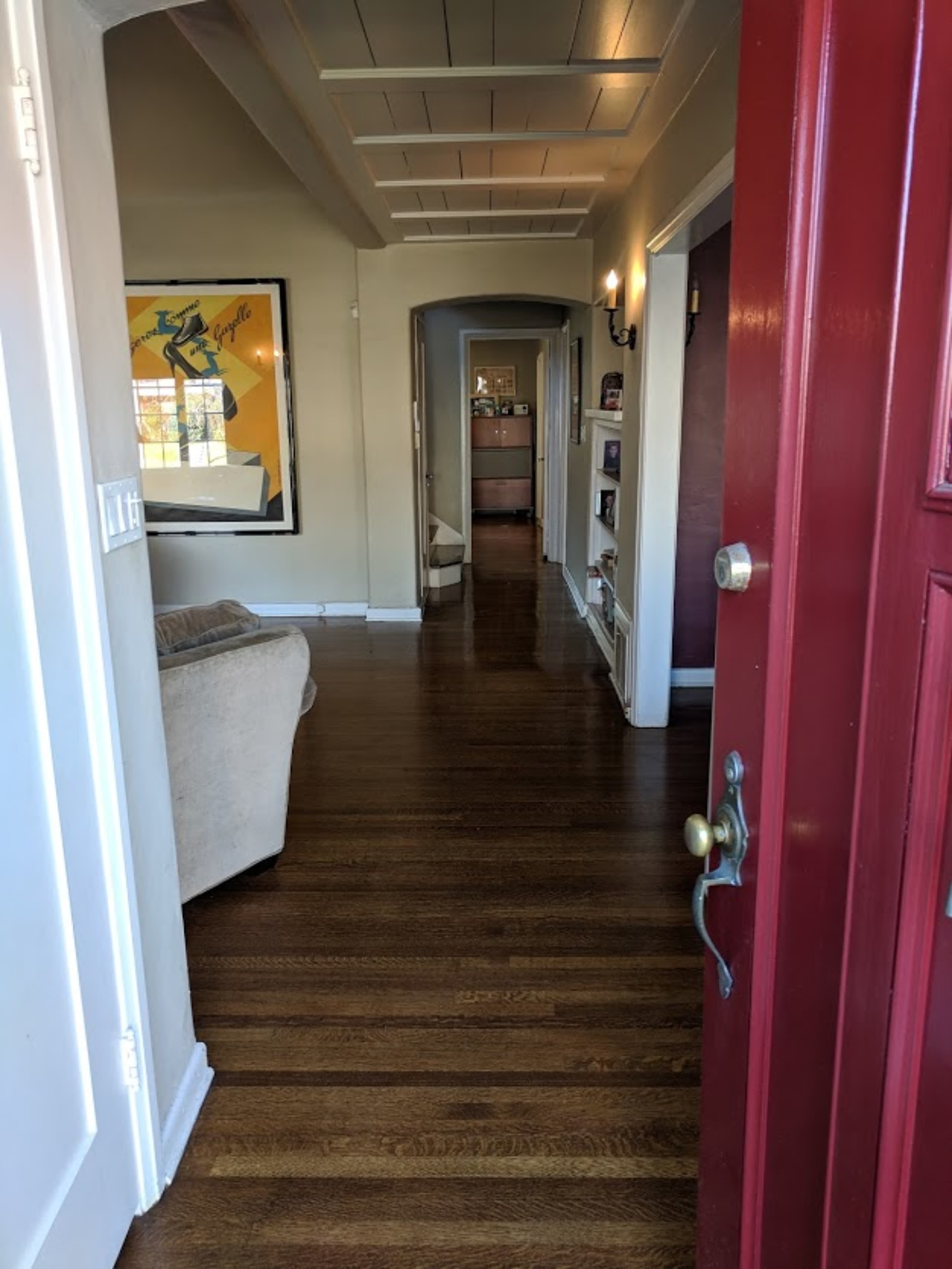 A view of a hallway from an open red front door, leading to a living area with wooden floors and white walls.