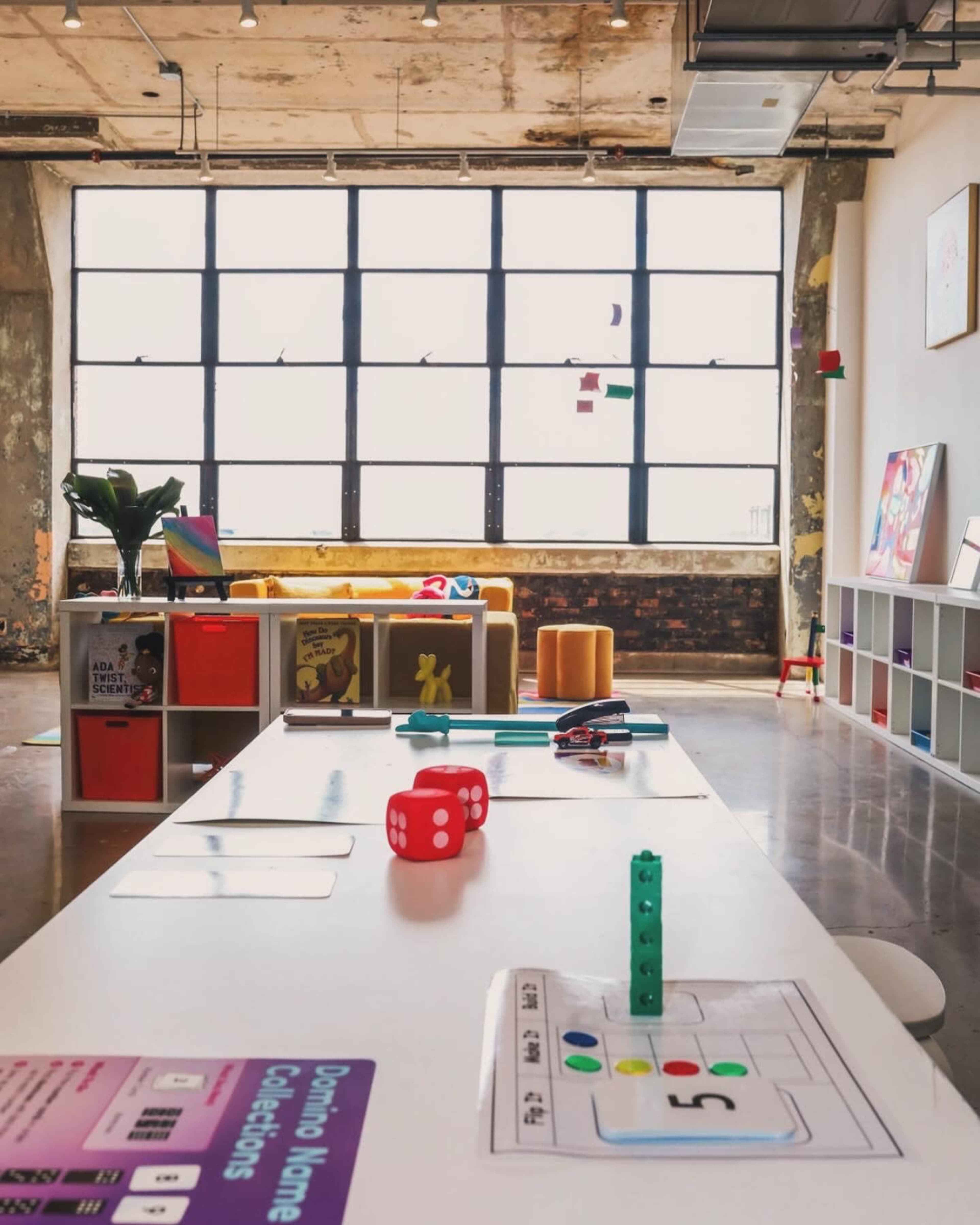 A brightly lit modern workspace with large windows, featuring a table set up with colored game pieces and educational materials, alongside a cozy seating area.
