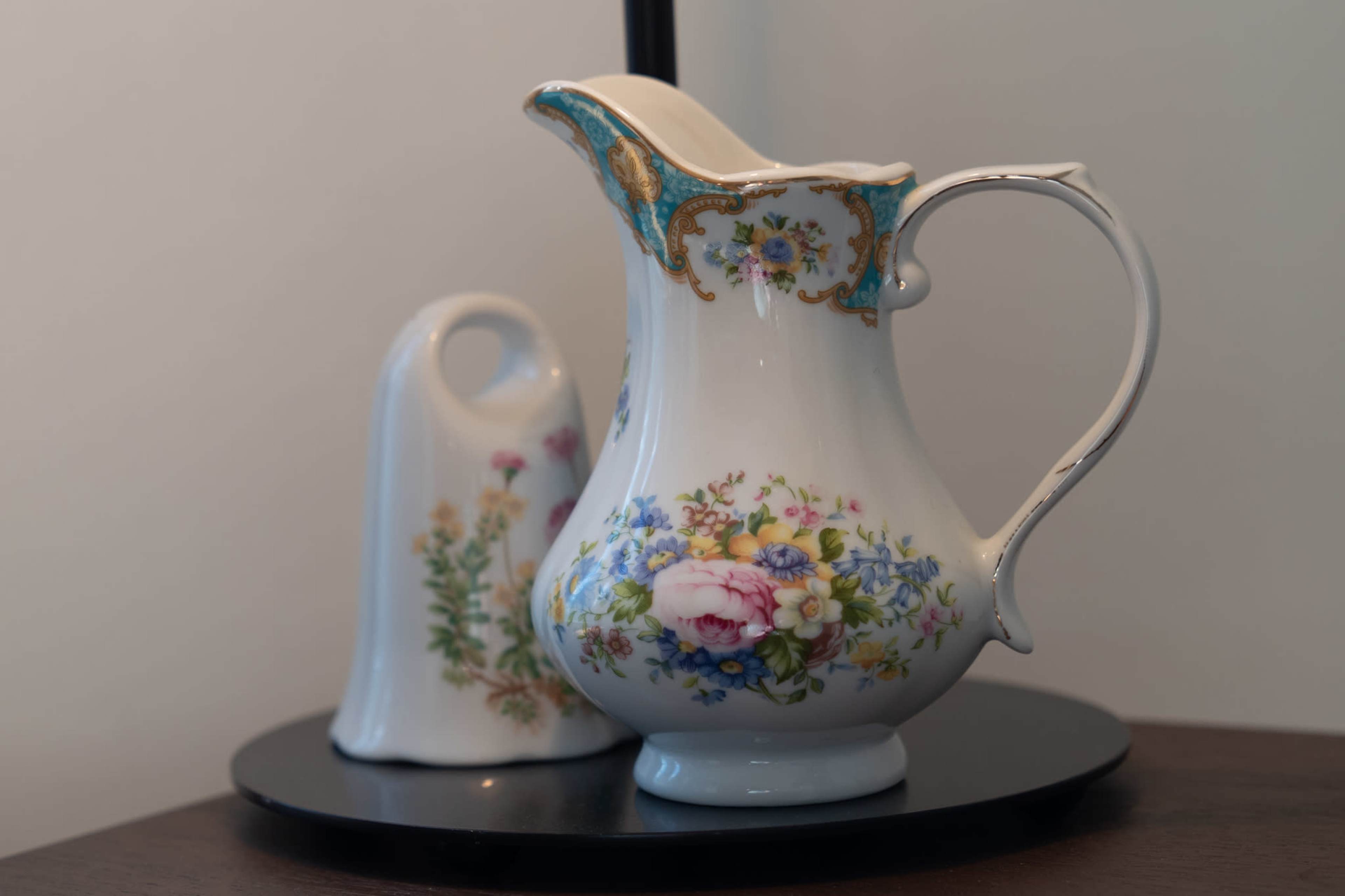 A floral-patterned porcelain pitcher alongside a small white porcelain bell, both placed on a round black tray.
