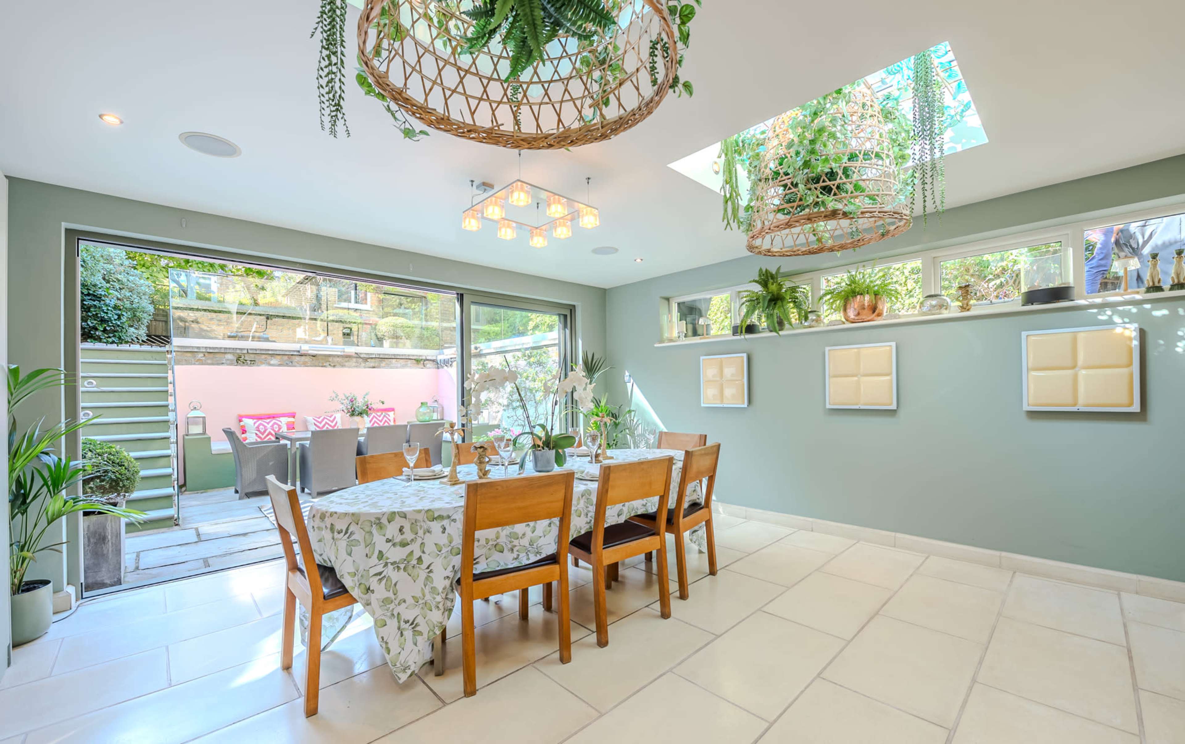 A modern dining area features a large table surrounded by wooden chairs, with large glass doors opening to an outdoor space and plants hanging from the ceiling.