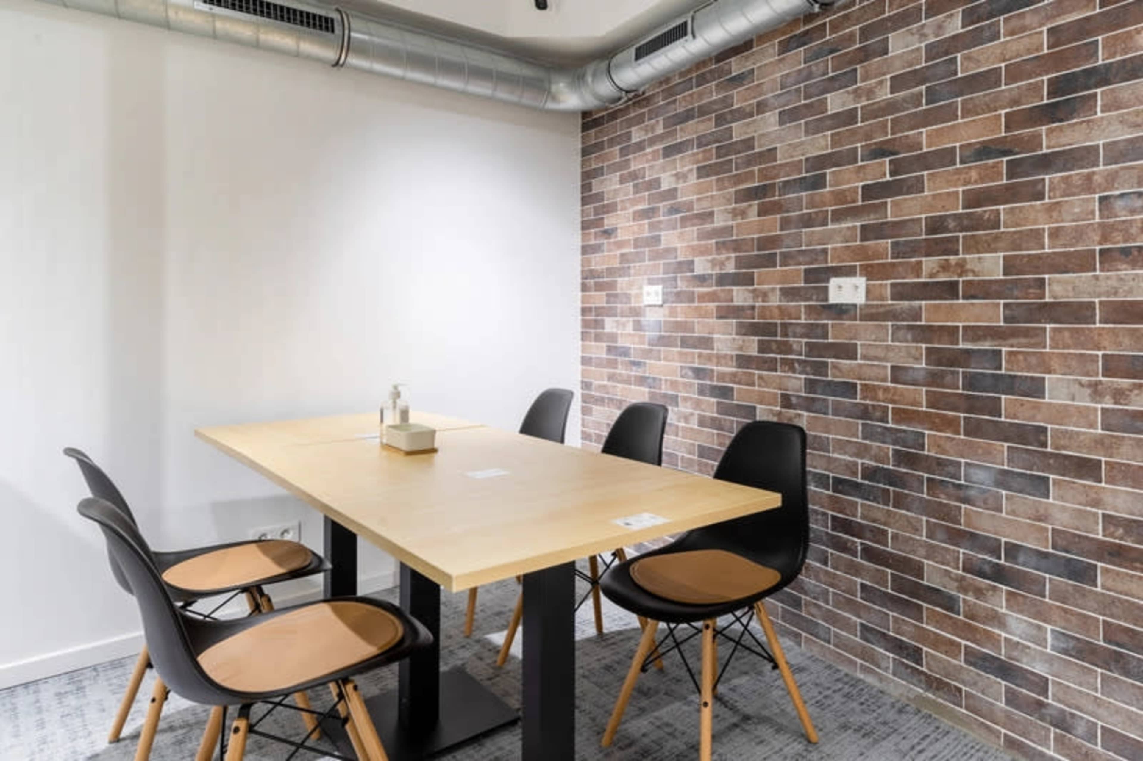 A simple meeting room features a wooden table surrounded by four black chairs, with a brick-patterned wall in the background.