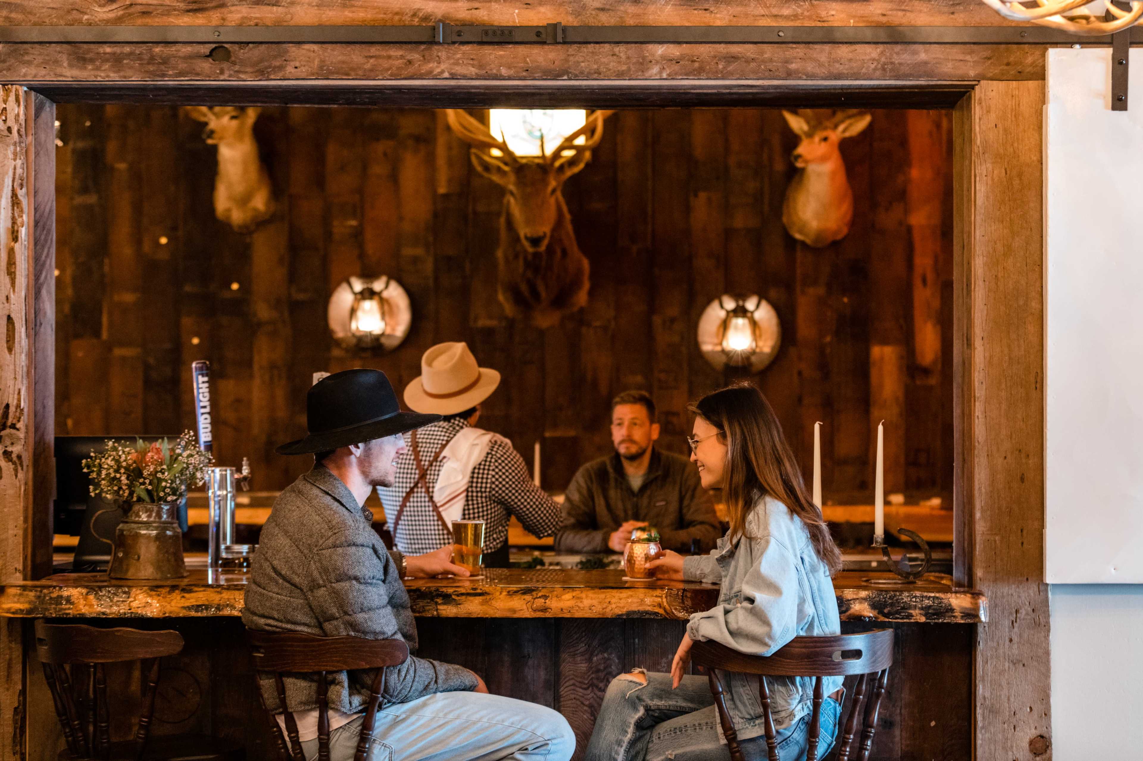 Two people sit at a bar engaged in conversation, while another person stands nearby and a fourth person sits in the background, with mounted deer heads adorning the wall.
