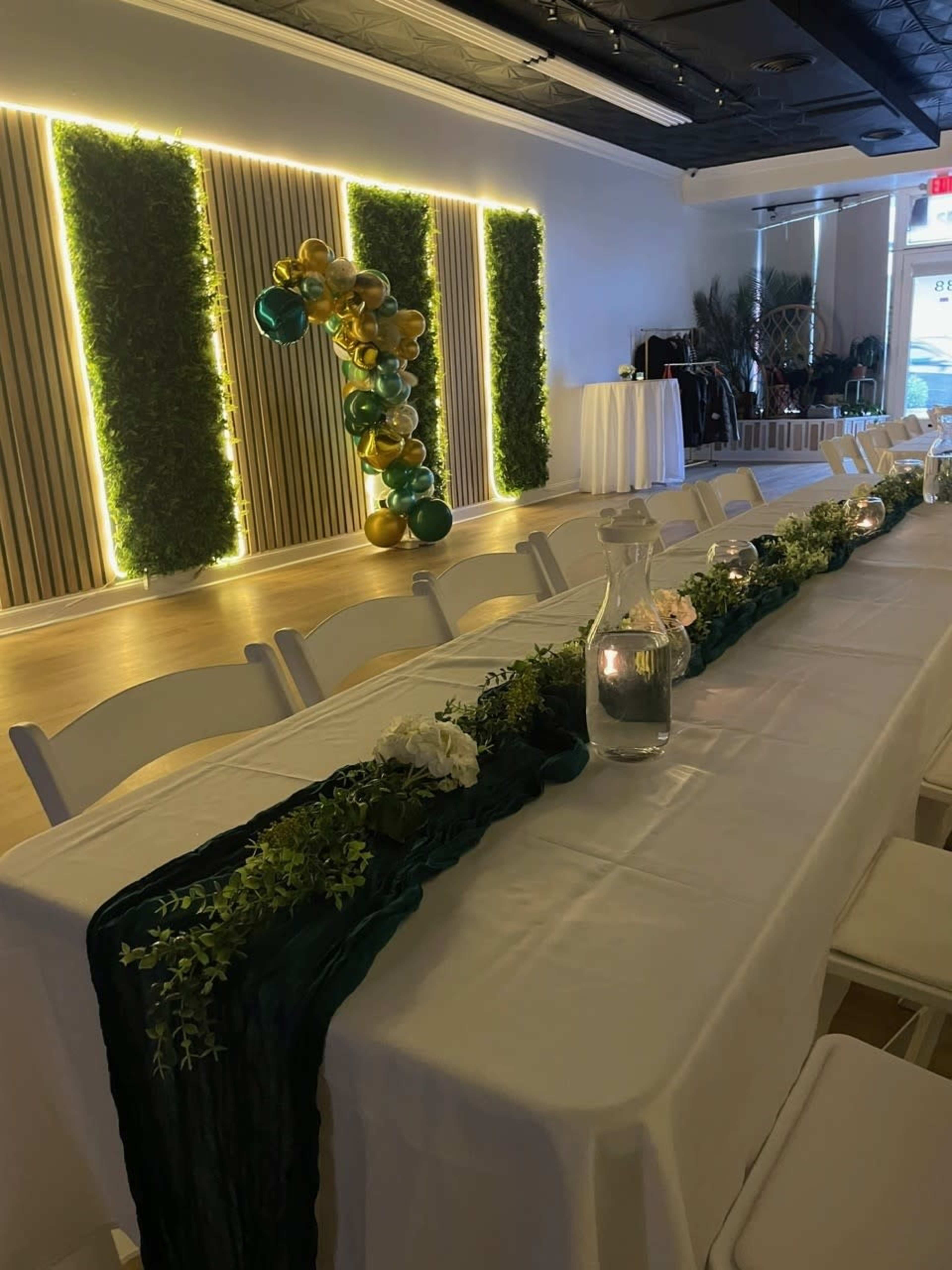 A long, elegantly decorated table with a green runner and floral arrangements is set up in a well-lit event space featuring plants and balloon decor on the walls.