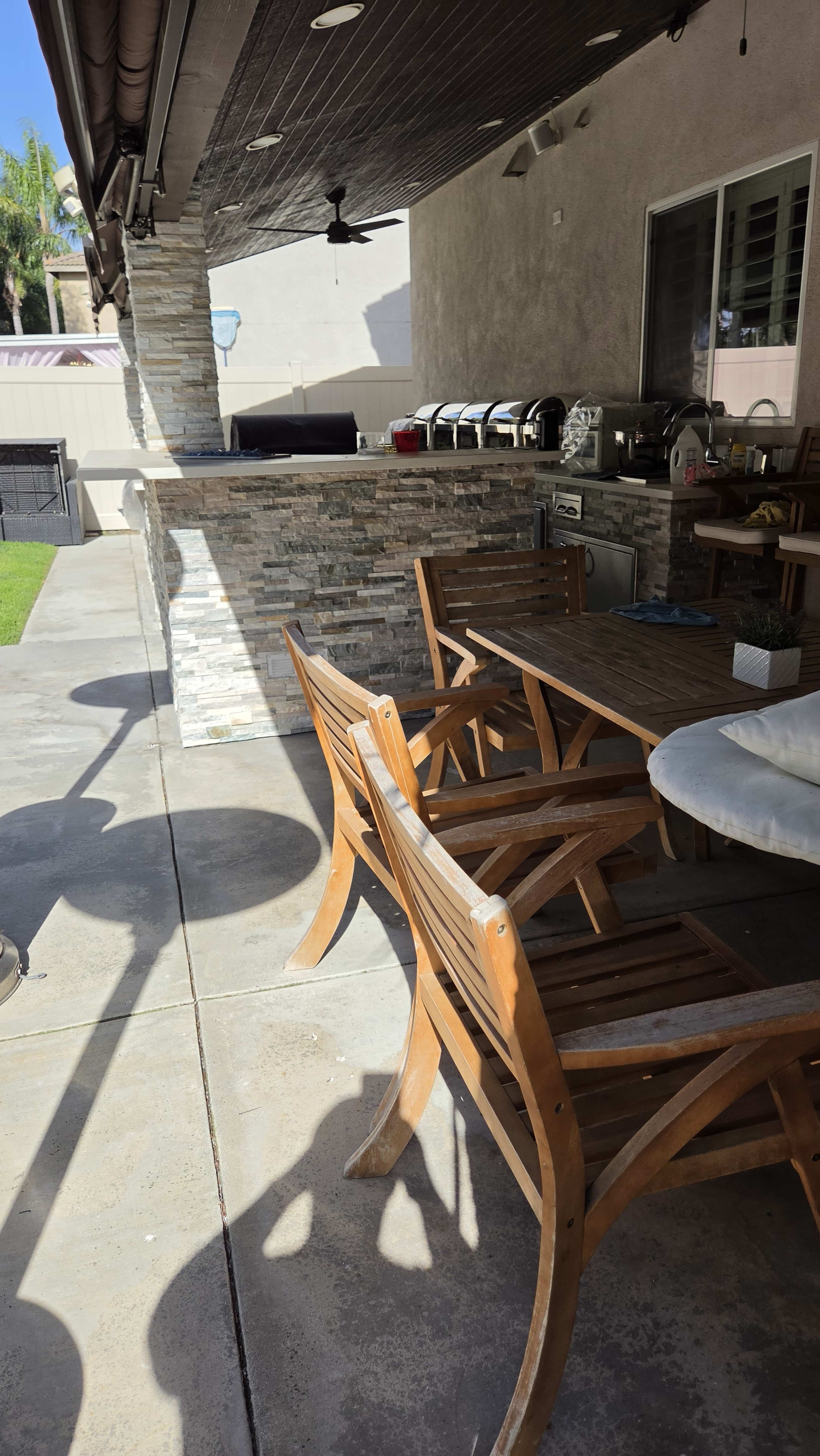 The image shows a patio area featuring a stone grill station, wooden dining furniture, and a concrete floor.