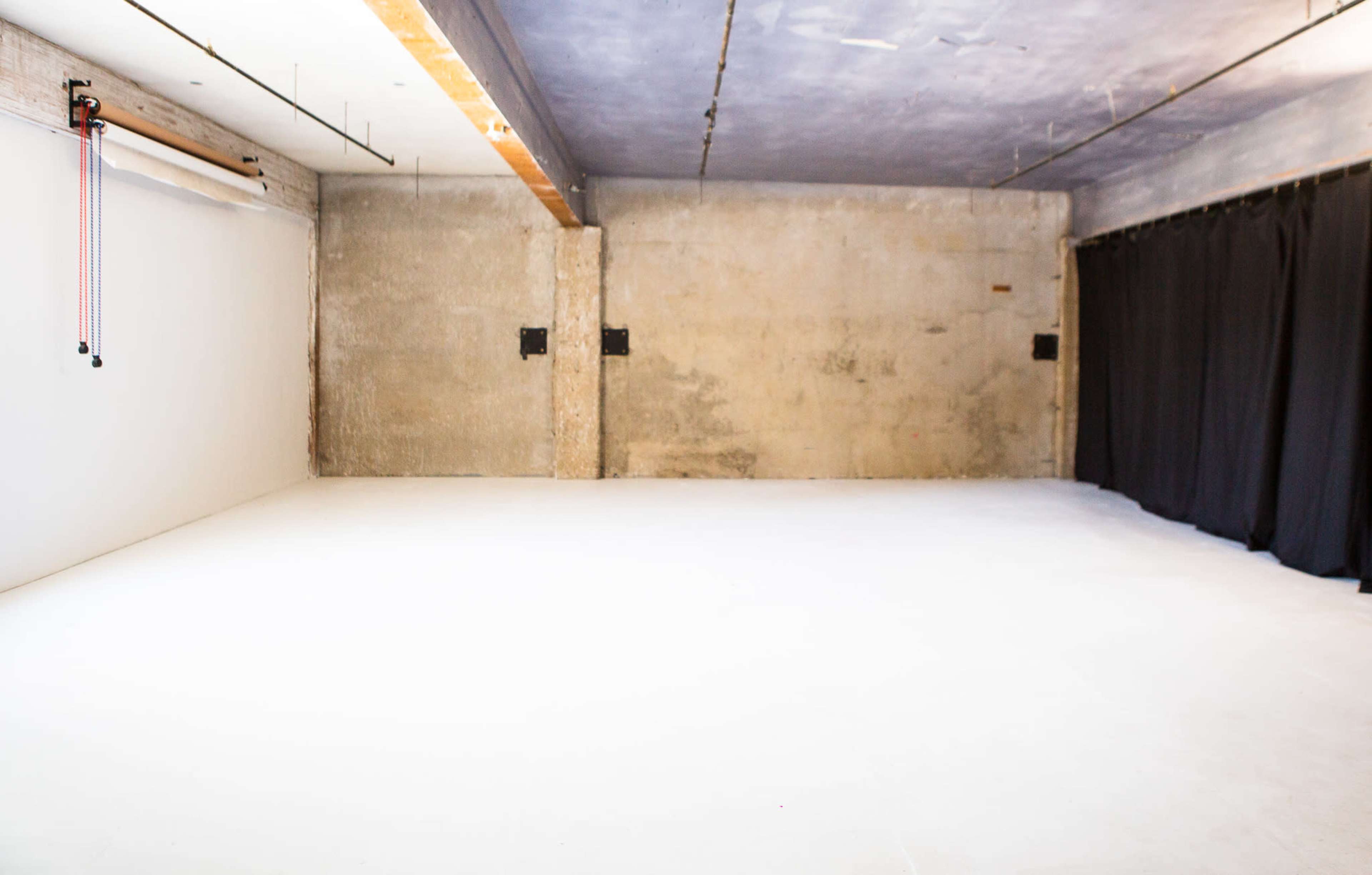 A spacious, empty studio with concrete walls and a smooth white floor, featuring a black curtain on one side.