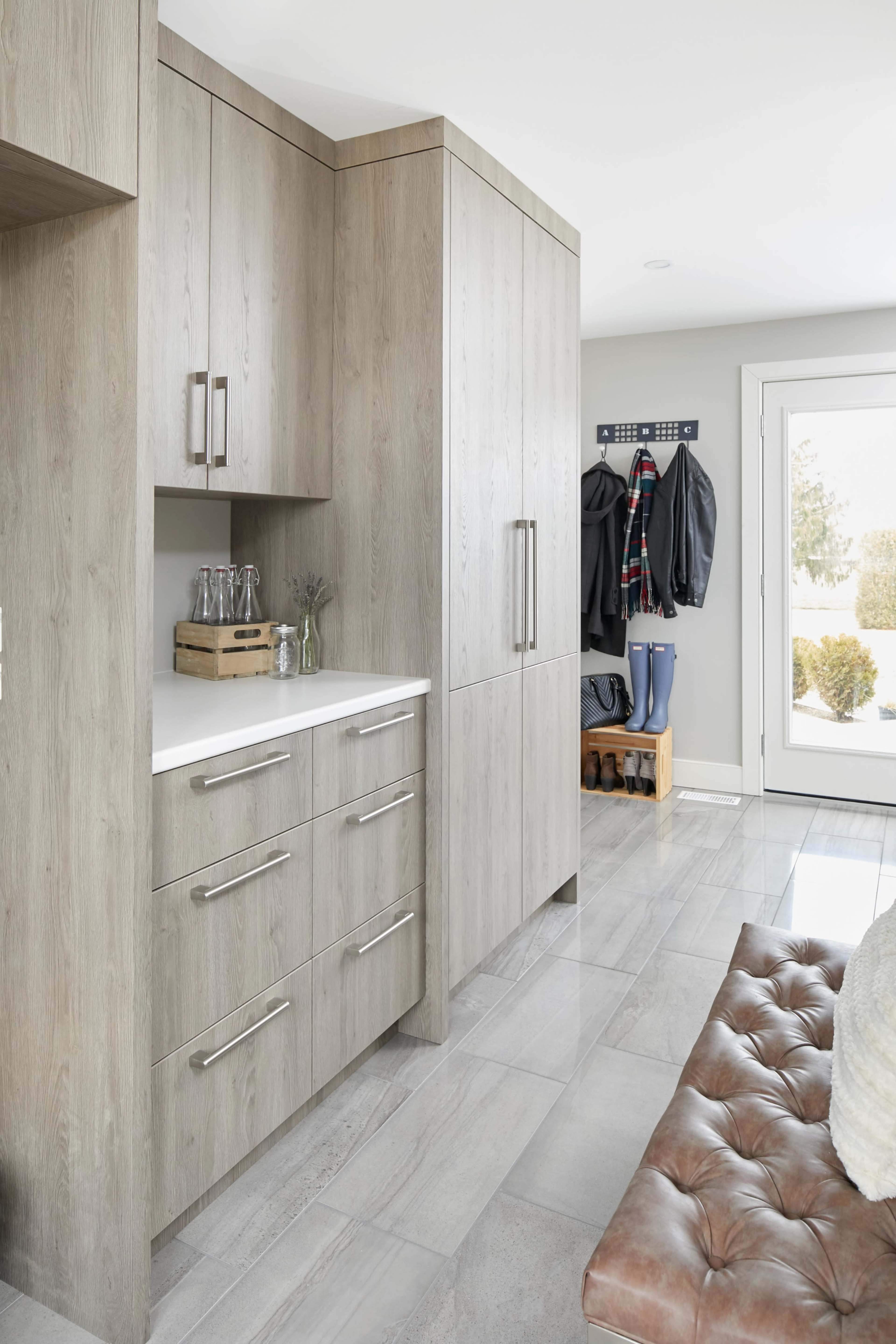 A modern entryway featuring cabinetry with a light wood finish, a countertop, and wall hooks for coats, alongside a bench and a window.
