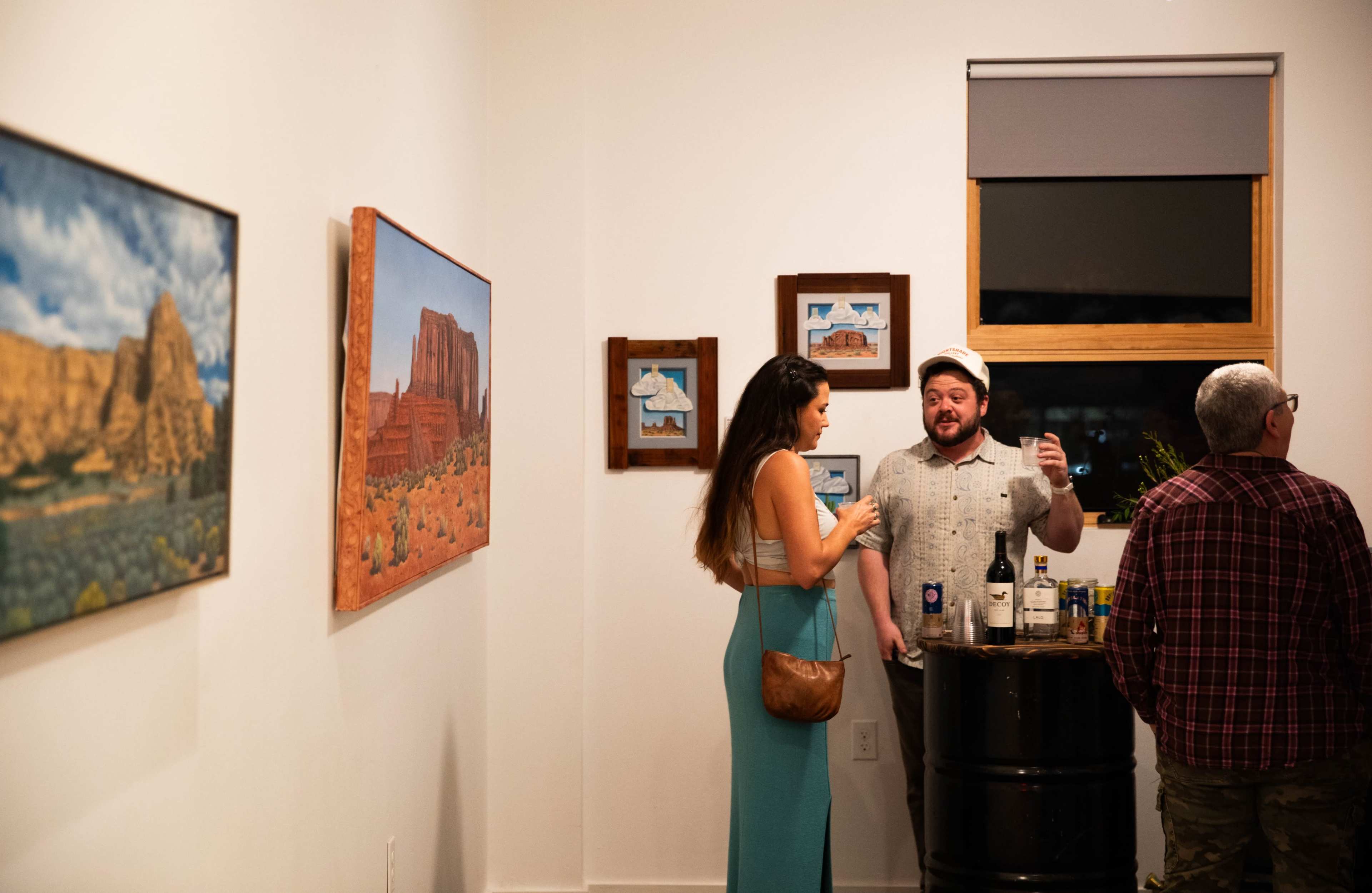 Two people are engaged in conversation while standing in an art gallery featuring landscape paintings on the walls.