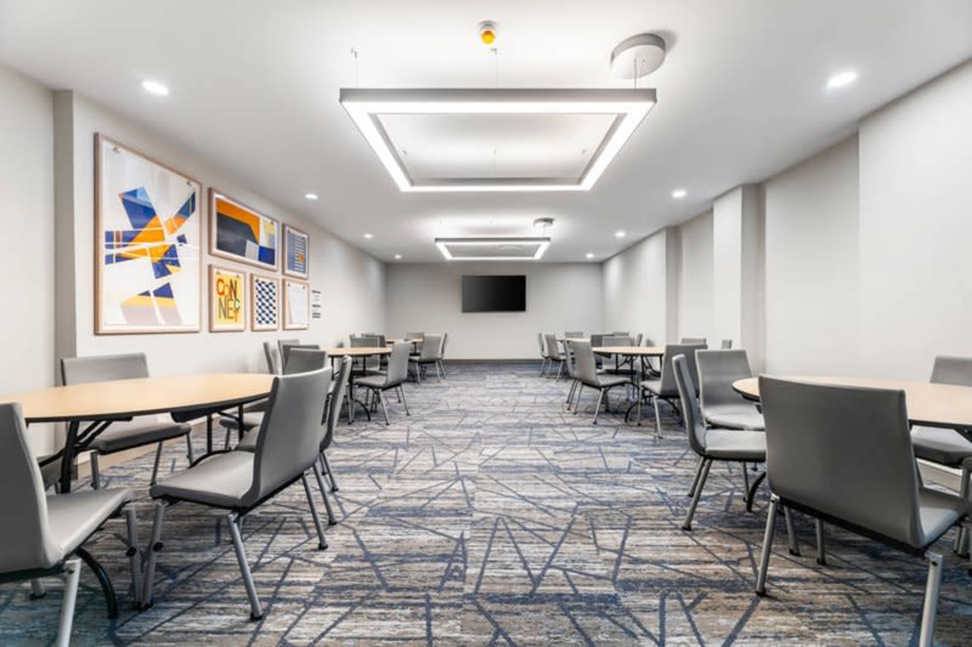The image shows a well-lit conference room with several round tables and modern chairs, accompanied by artwork on the walls and a television screen.