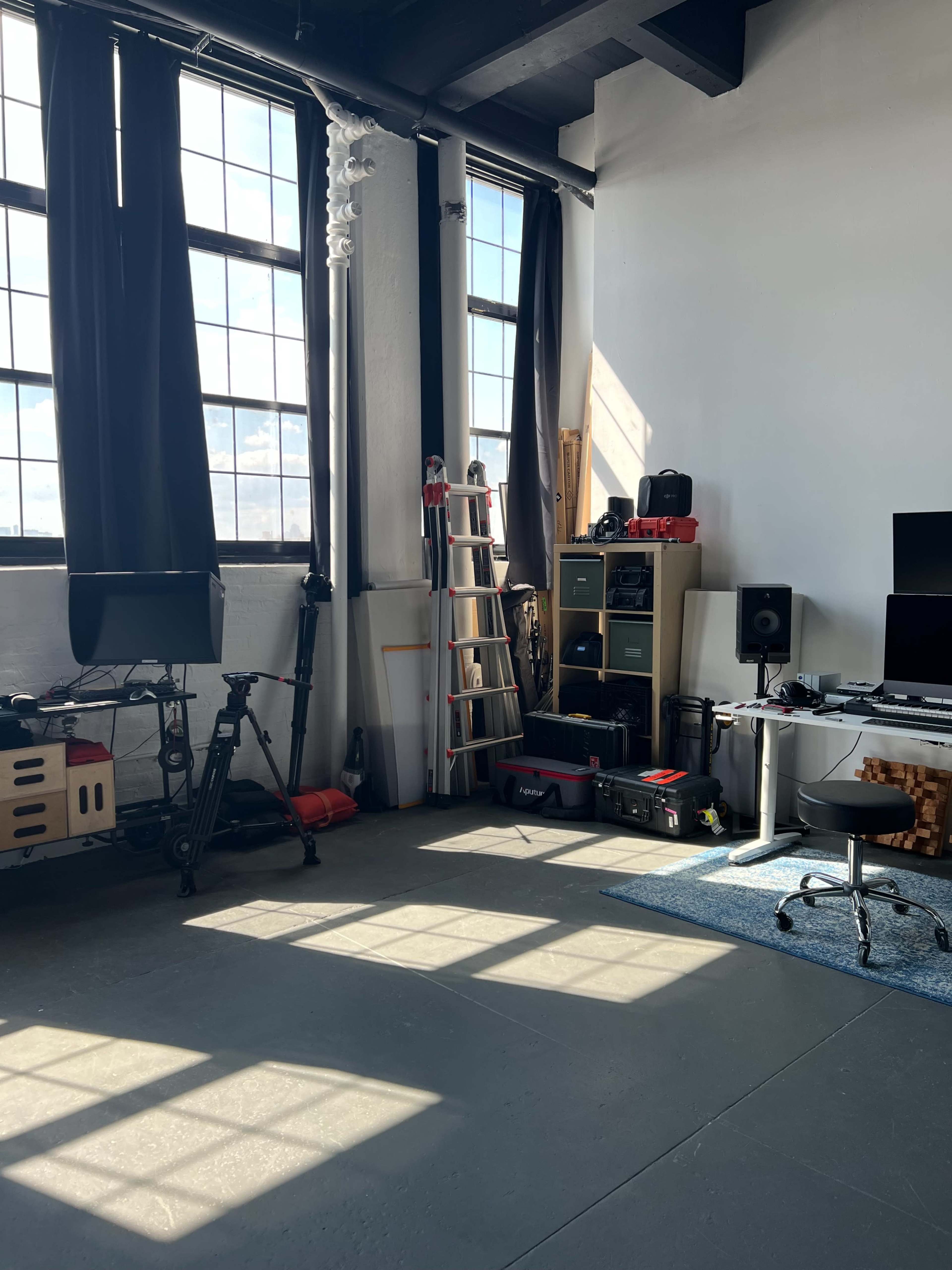 An industrial-style room with large windows, a ladder, various equipment, and a workspace setup.