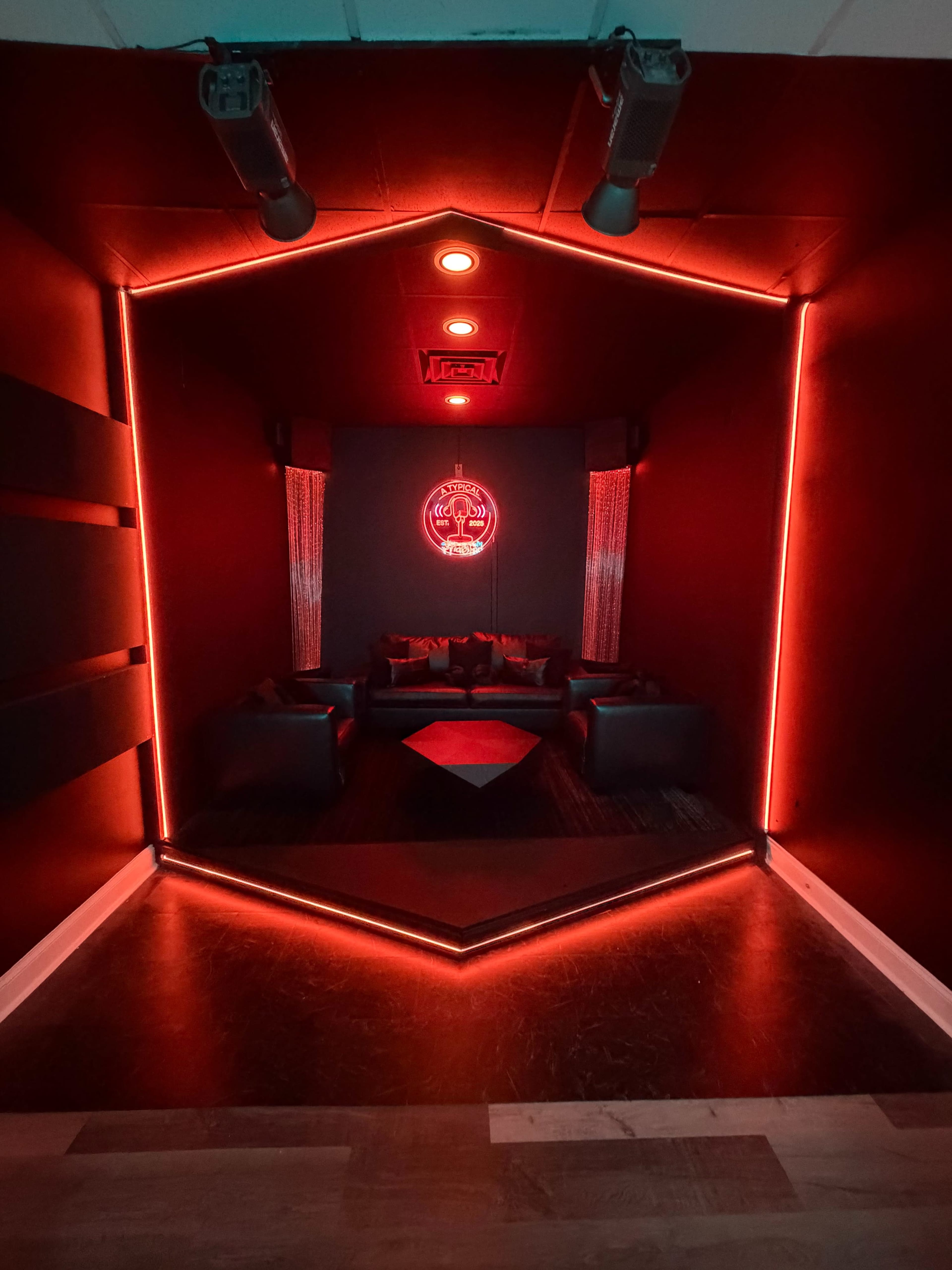 The image shows a modern room with black walls, dim lighting, and red neon accents, featuring a seating area with dark couches arranged around a central hexagonal table.
