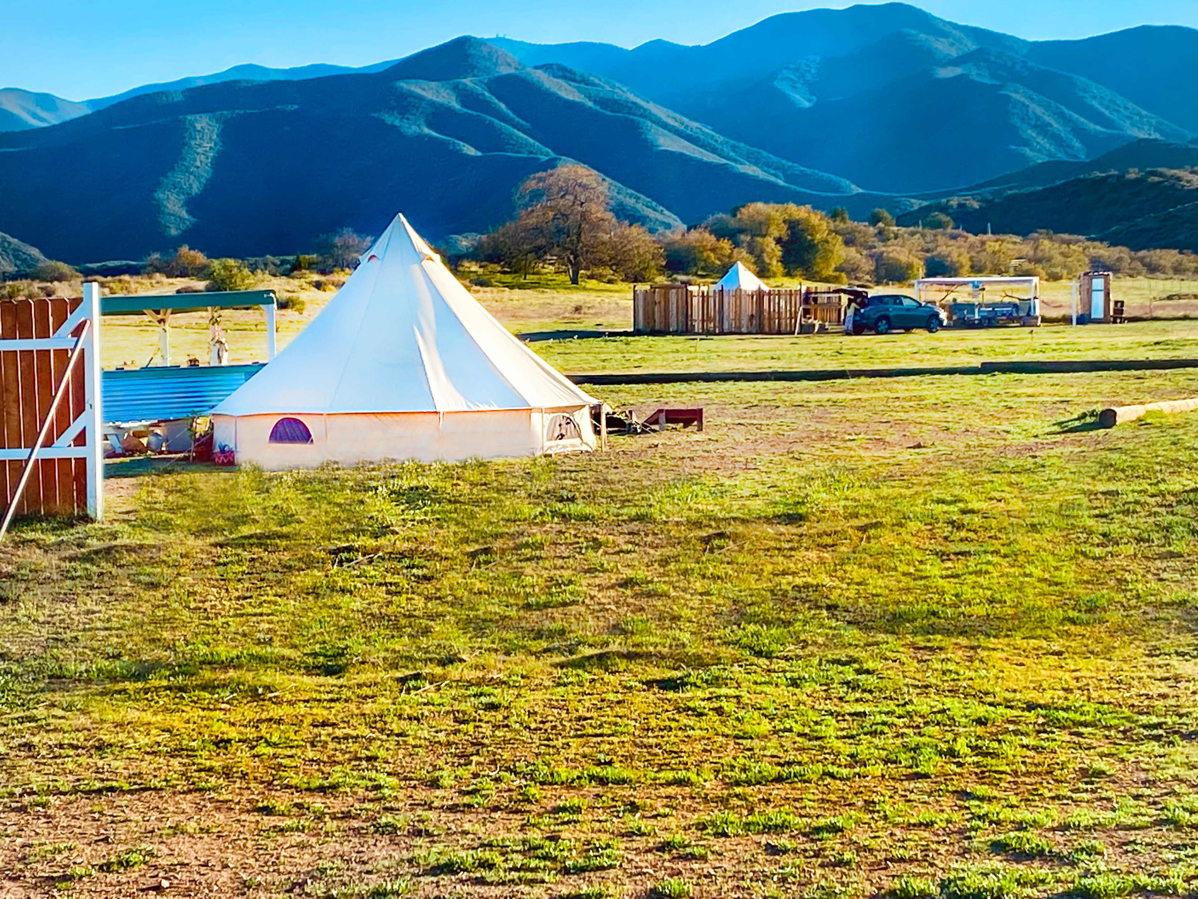 A white tent is set up in a grassy area with mountains in the background, along with a vehicle and additional structures nearby.