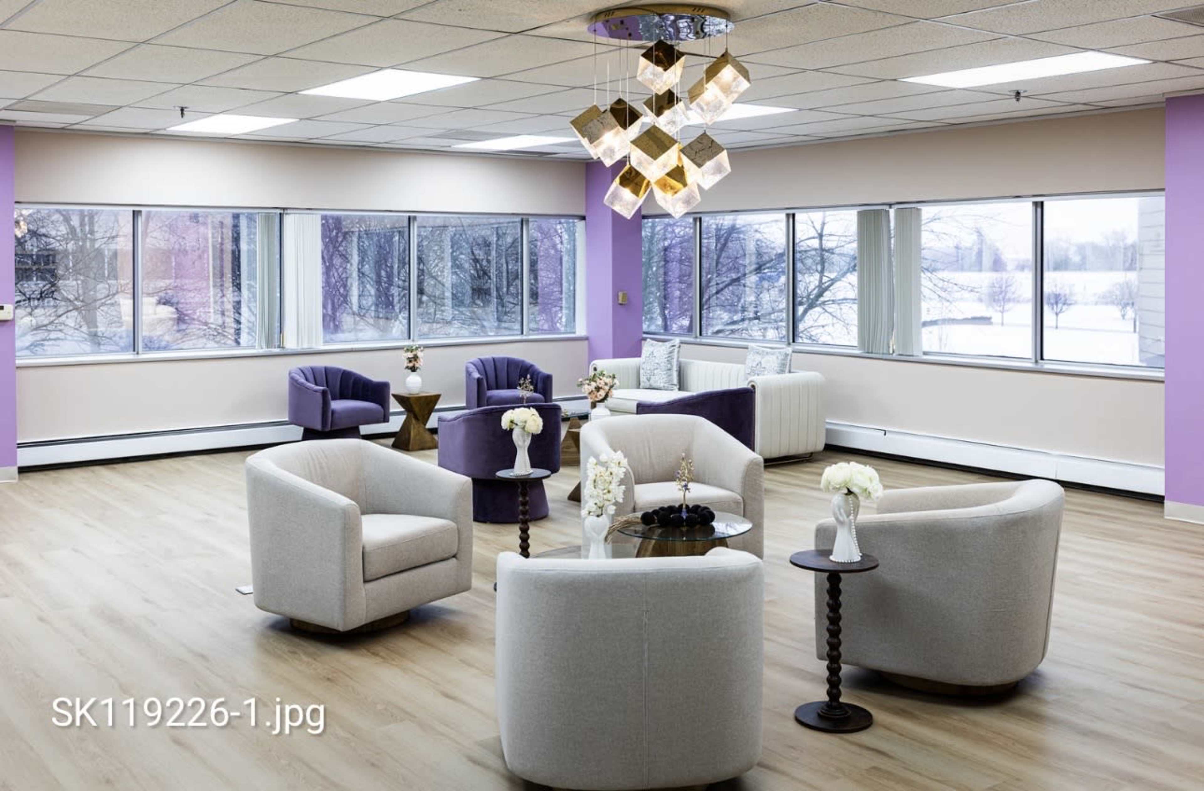 The image depicts a modern lounge area with several white and purple seating arrangements, a coffee table, and large windows allowing natural light to fill the space.