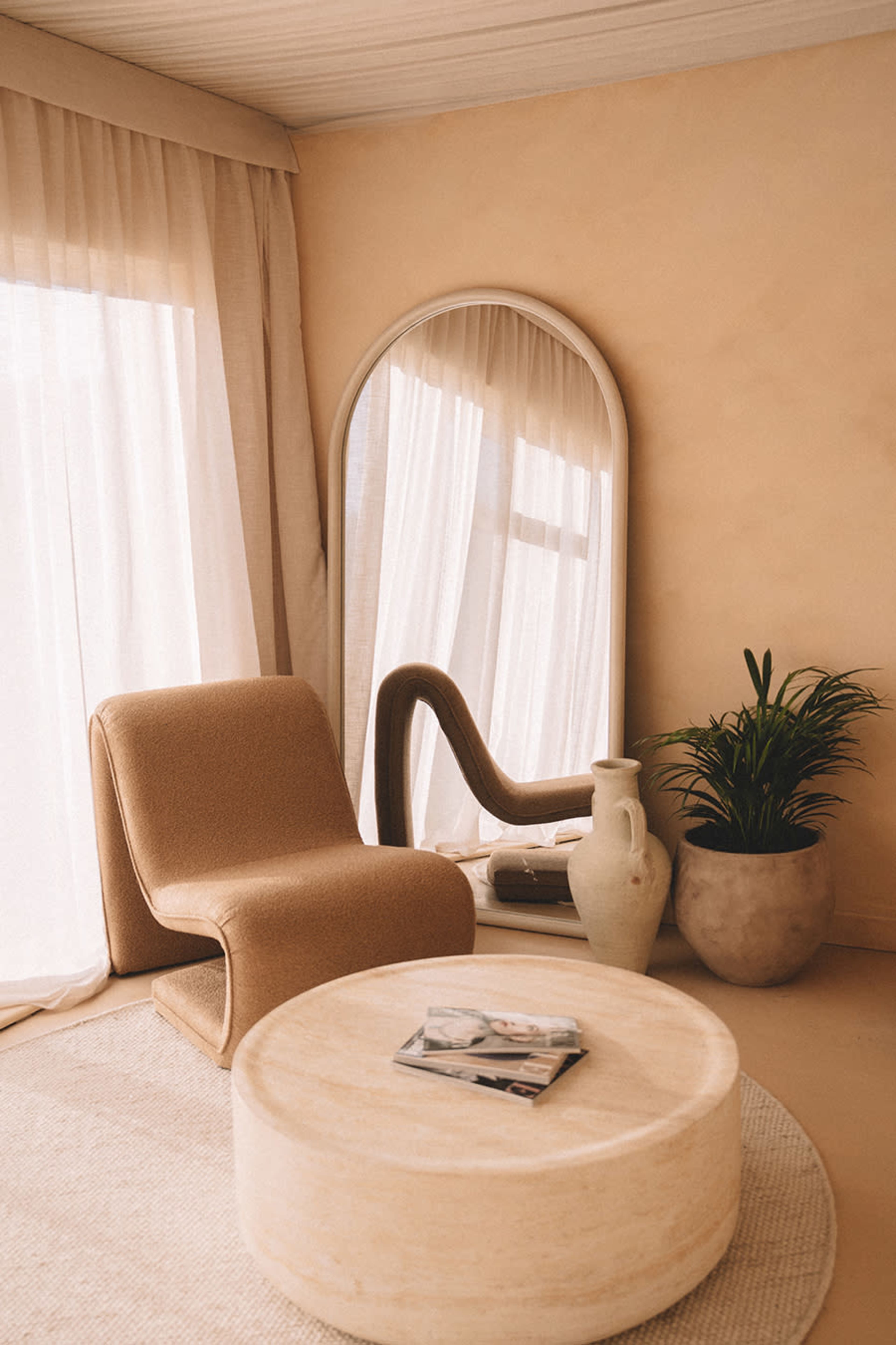 The image shows a minimalist living space featuring a curved chair, a round coffee table, a large mirror, and a potted plant, all set against light-colored walls and curtains.