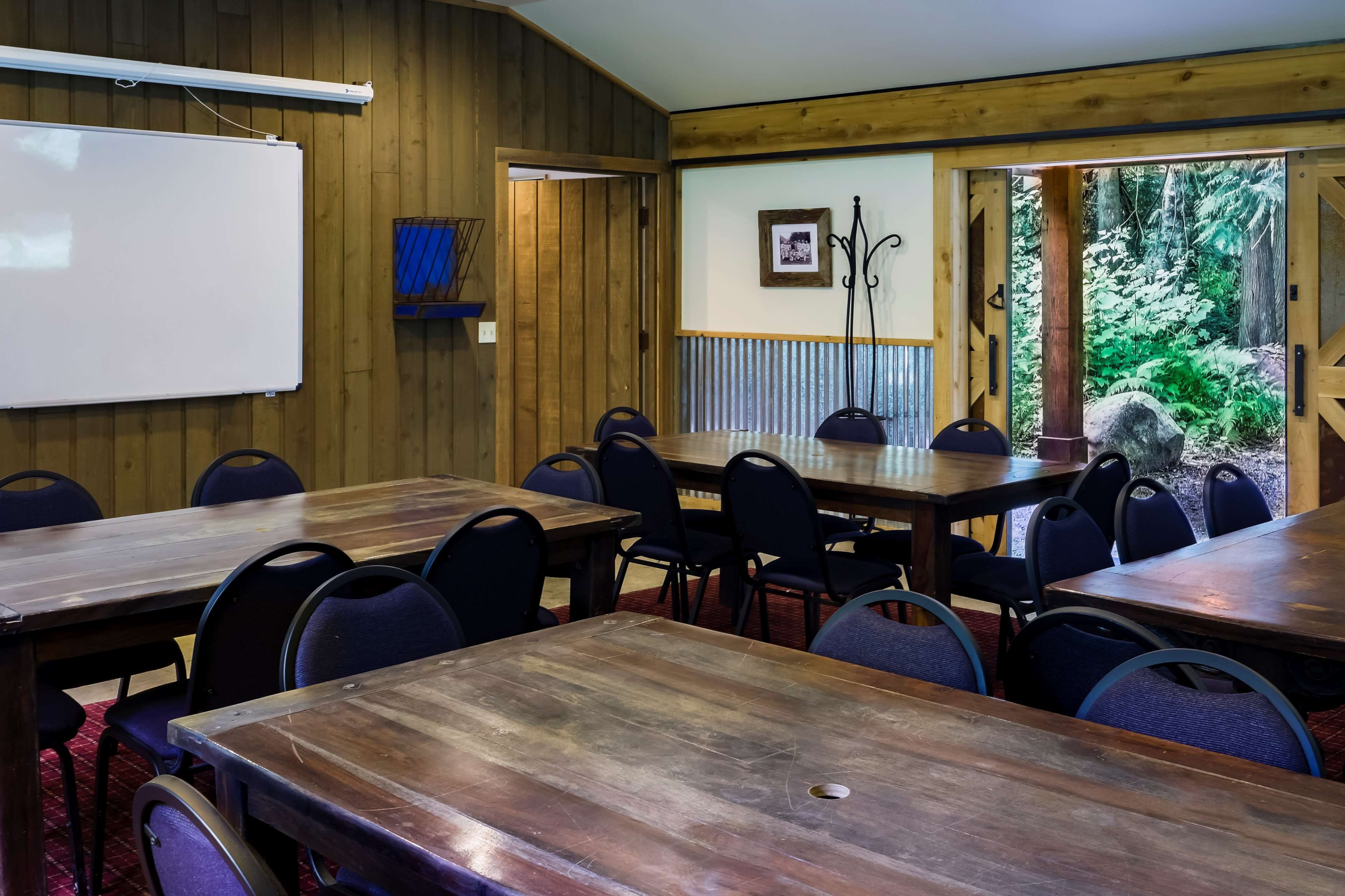 A rustic meeting room features several wooden tables arranged with black chairs, a whiteboard on the wall, and an open door leading to a forested area outside.