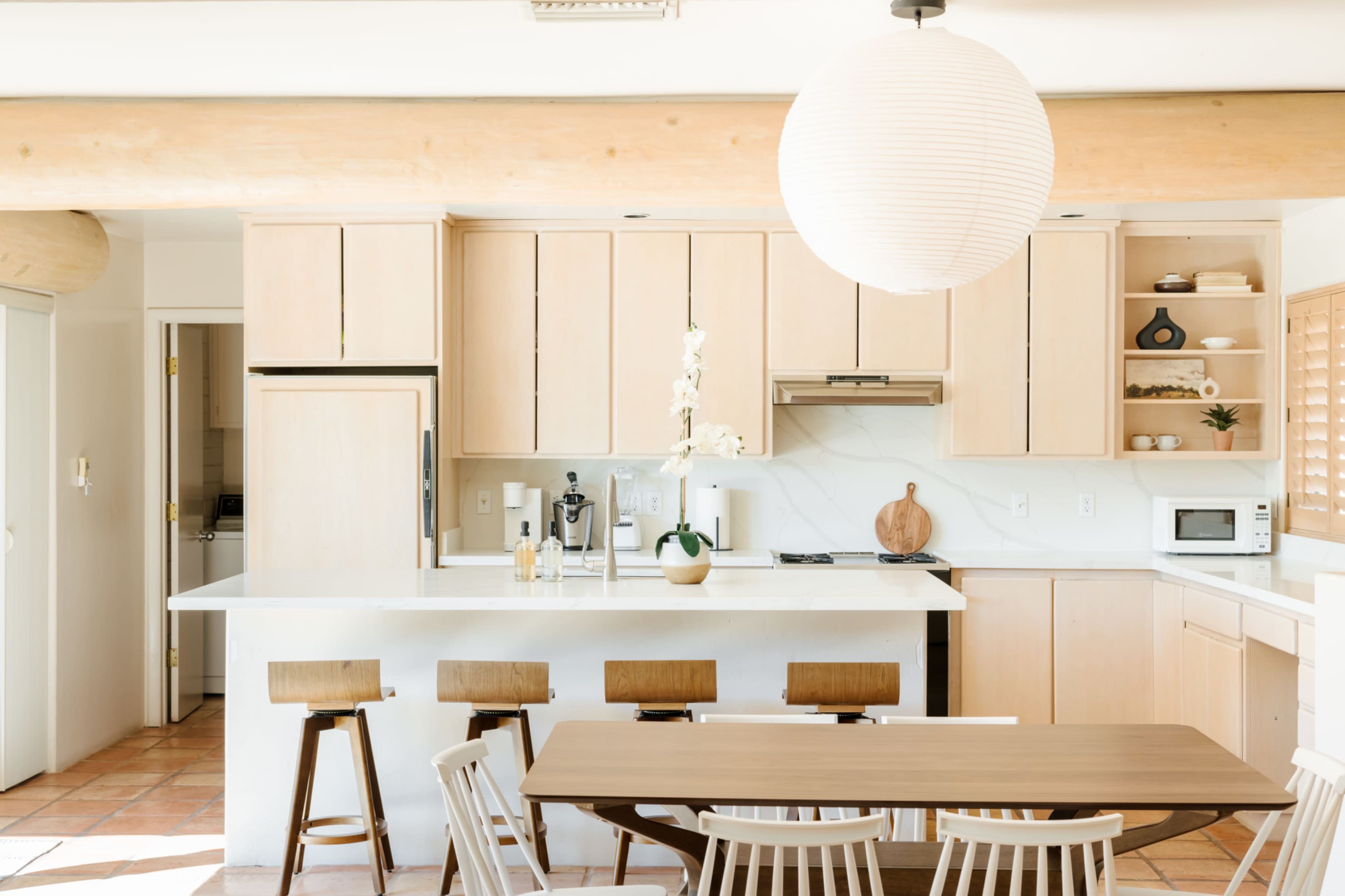 A modern kitchen features light wood cabinetry, a central island, and a dining table surrounded by white chairs.