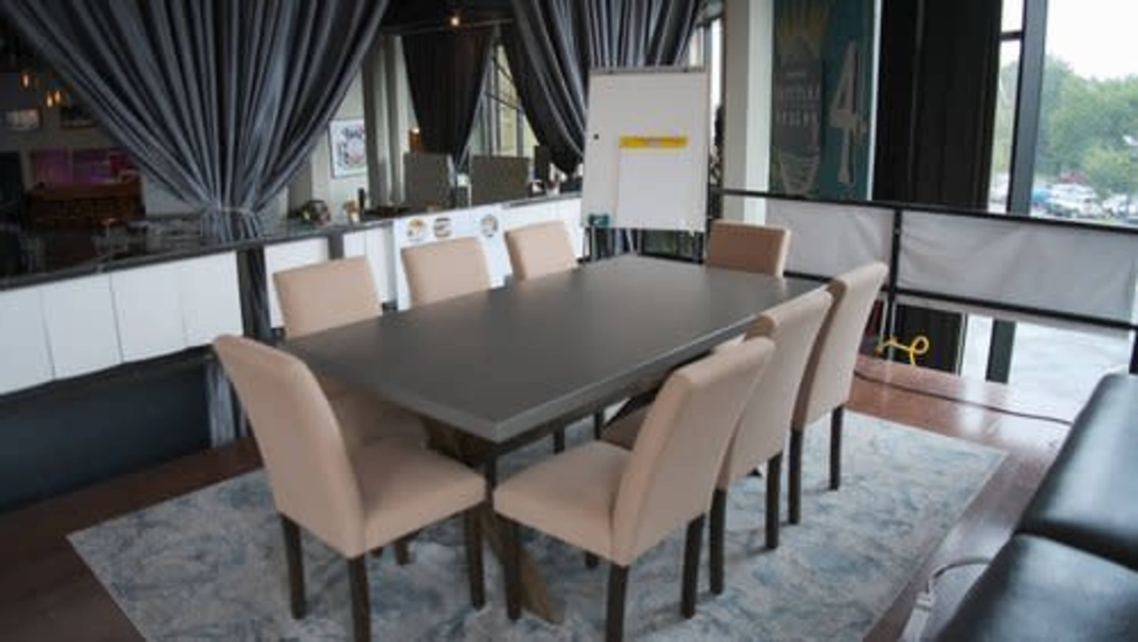 The image shows a conference room with a large rectangular table surrounded by eight beige chairs and a flip chart on an easel.