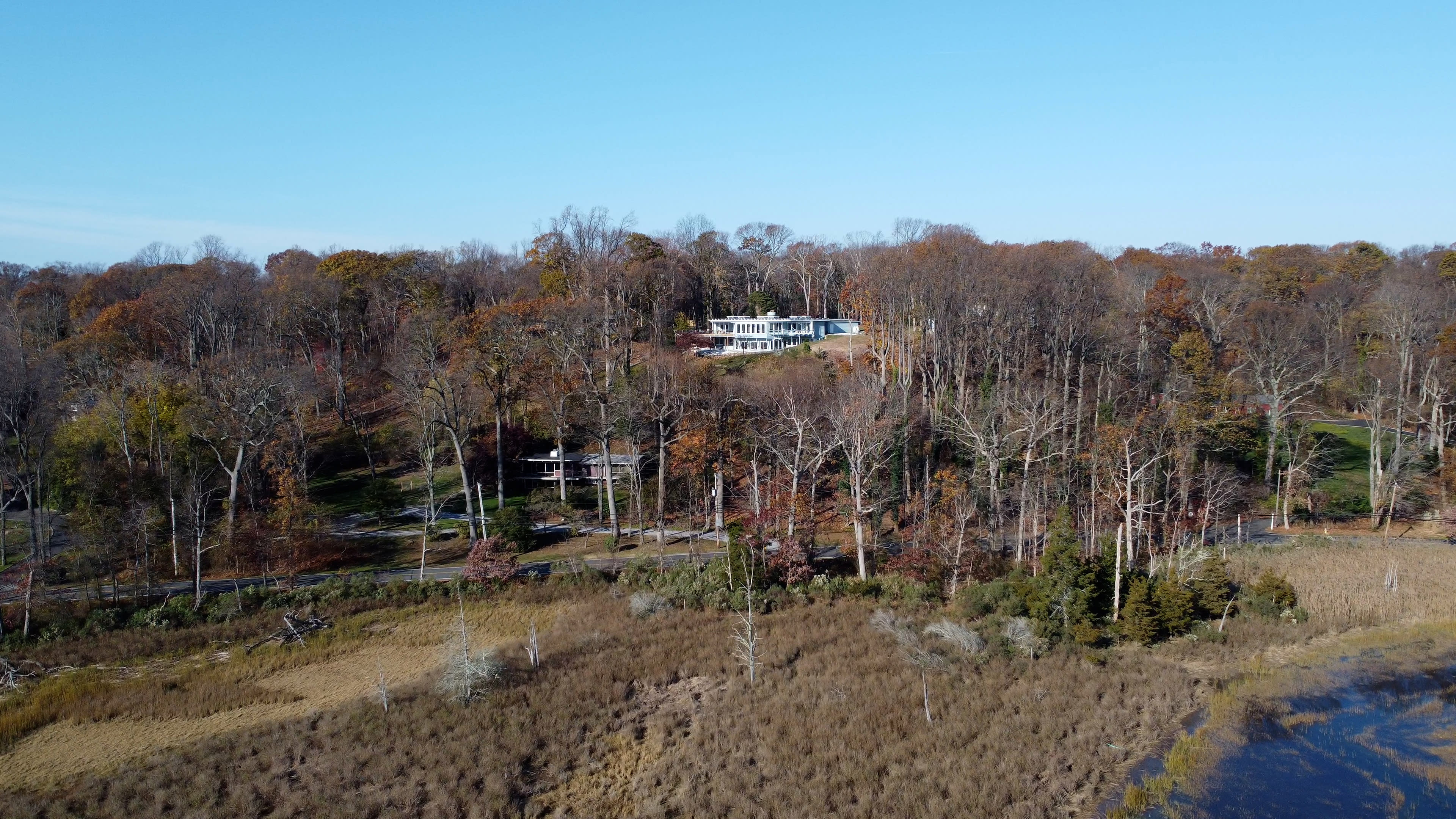 A modern house is situated on a hillside, surrounded by bare trees and autumn foliage near a water body.